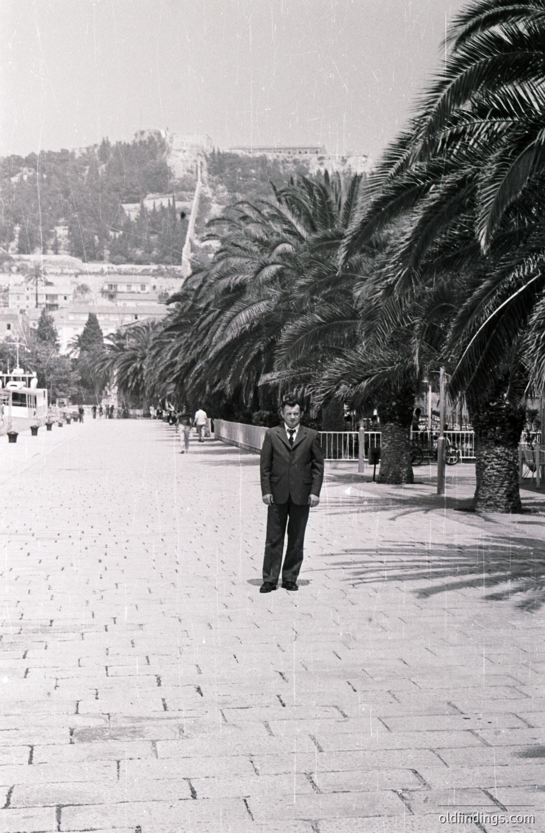 A formally dressed man stands on a patterned stone promenade lined with palm trees. A coastal cityscape rises in the distance, featuring tiered buildings and a visible harbor. Likely 1960s-1970s, possibly a seaside resort location. Commercial potential for design, travel imagery.