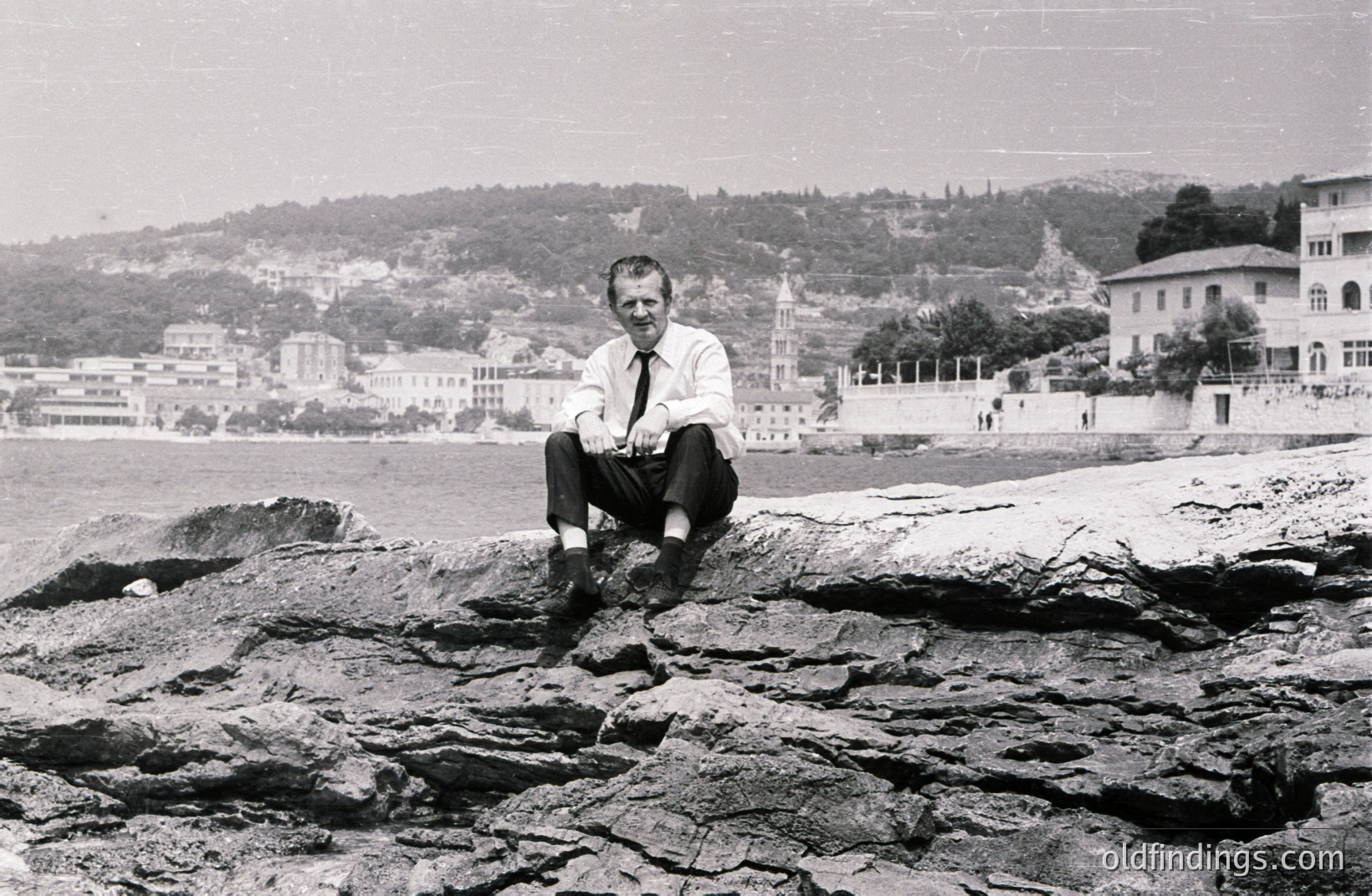 Man in a suit sits on rocks overlooking a seaside town and harbor. Appears to be a promotional portrait, perhaps for tourism or business. Architectural details suggest a Mediterranean location—likely early 1960s. Formal attire contrasts with the natural, rocky setting. A timeless image with potential for design applications.