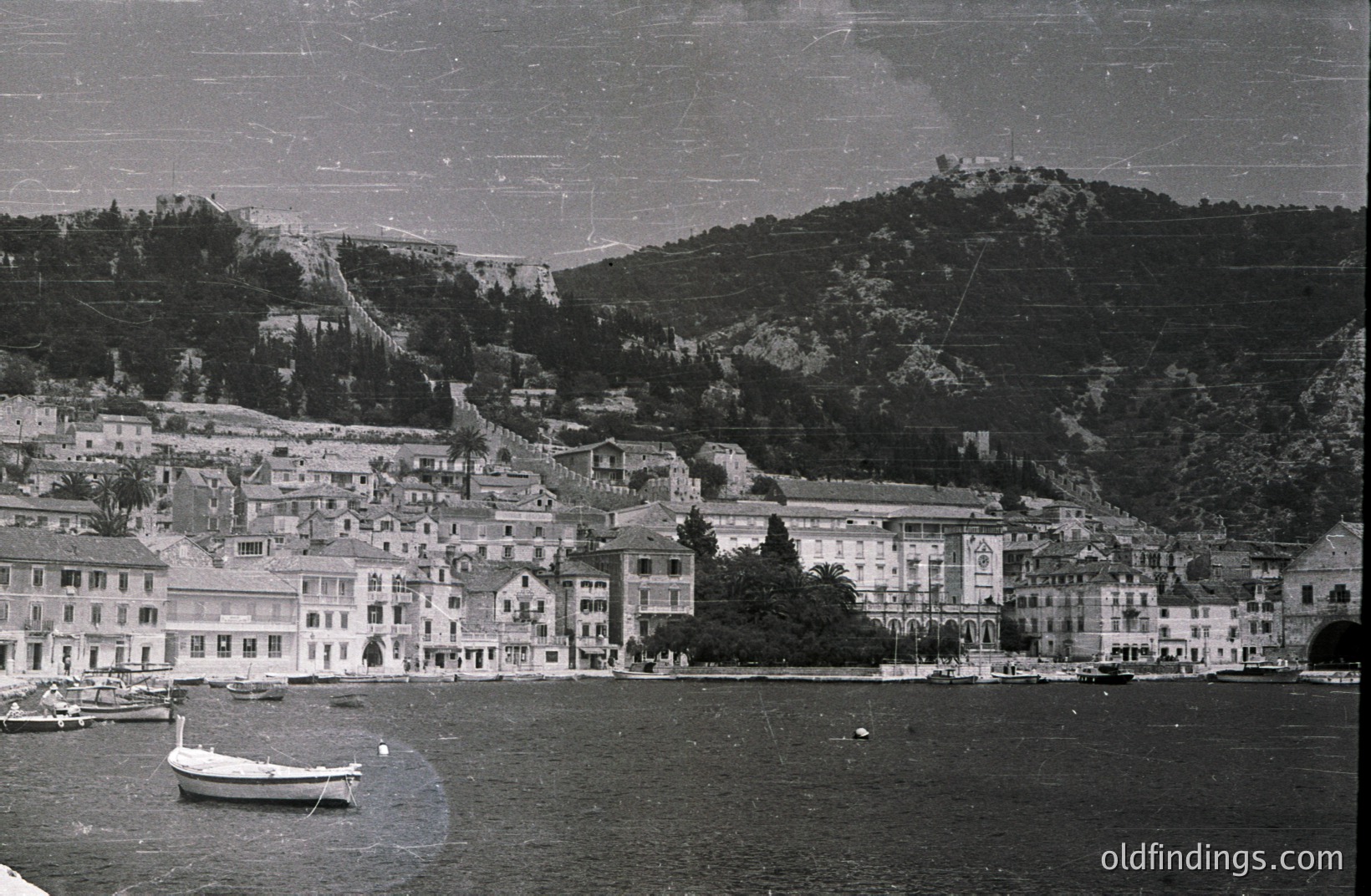 Seaside townscape with tiered architecture, likely Adriatic region. Buildings exhibit classical detailing & balconies. A small boat rests on the shore, facing a backdrop of forested hills & a fortress on a peak. Appears to be a tourist destination.