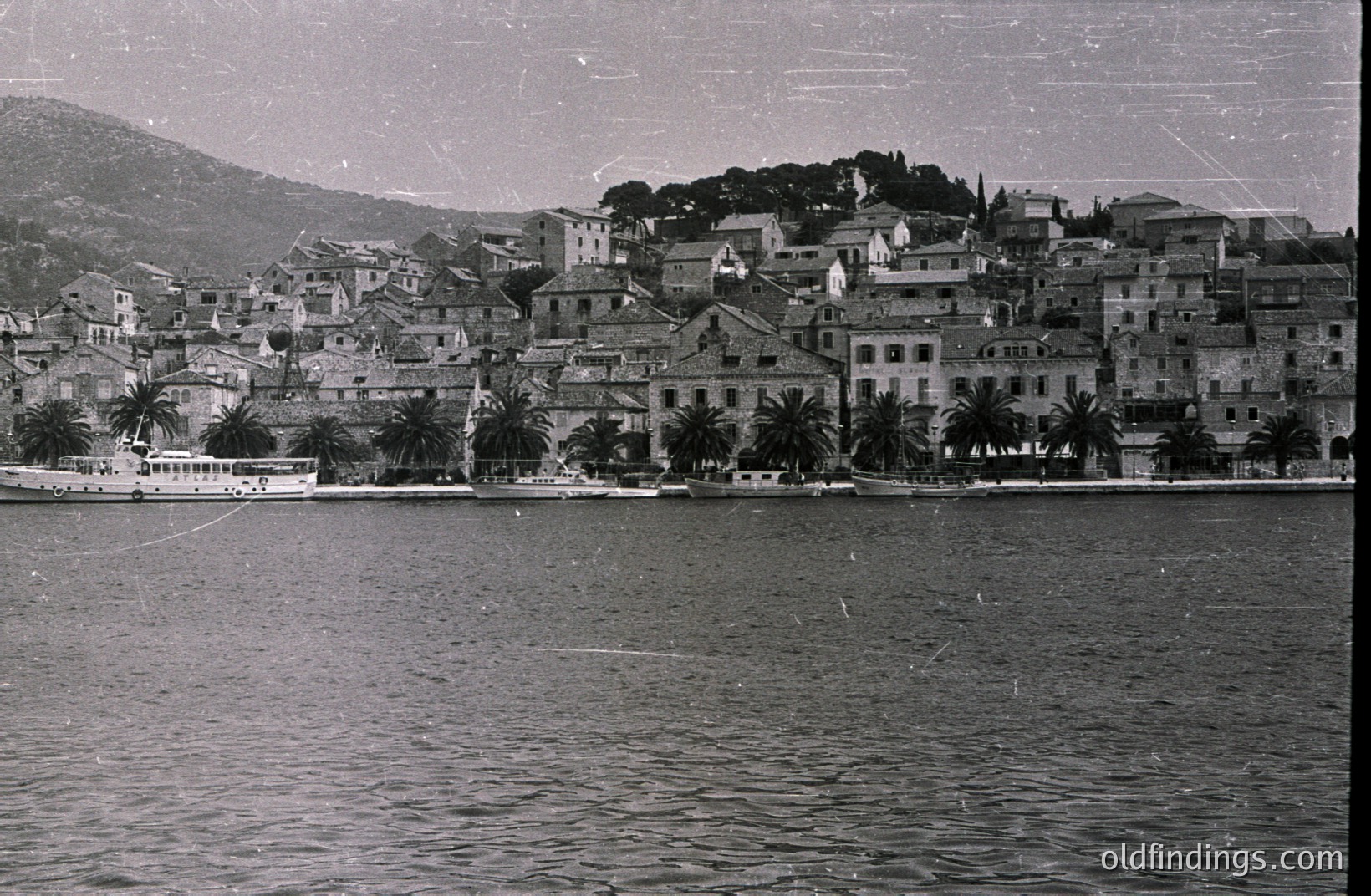 Coastal town scene, likely Mediterranean, with tightly packed buildings rising up a hillside. A small boat is docked along the waterfront. Palm trees are interspersed among the structures. Appears to be a vintage black and white photograph, potentially 1950s-1970s.