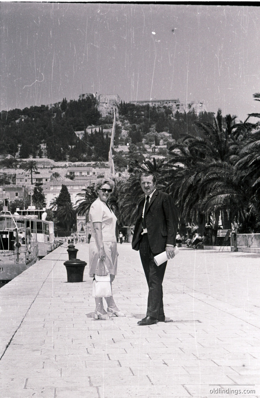 A formally dressed couple poses on a stone promenade, likely during a vacation. The woman wears a sheath dress and carries a handbag; the man sports a suit and tie, holding documents. A steep, terraced hillside with buildings rises behind them, featuring palm trees. Likely 1960s/70s travel photography.