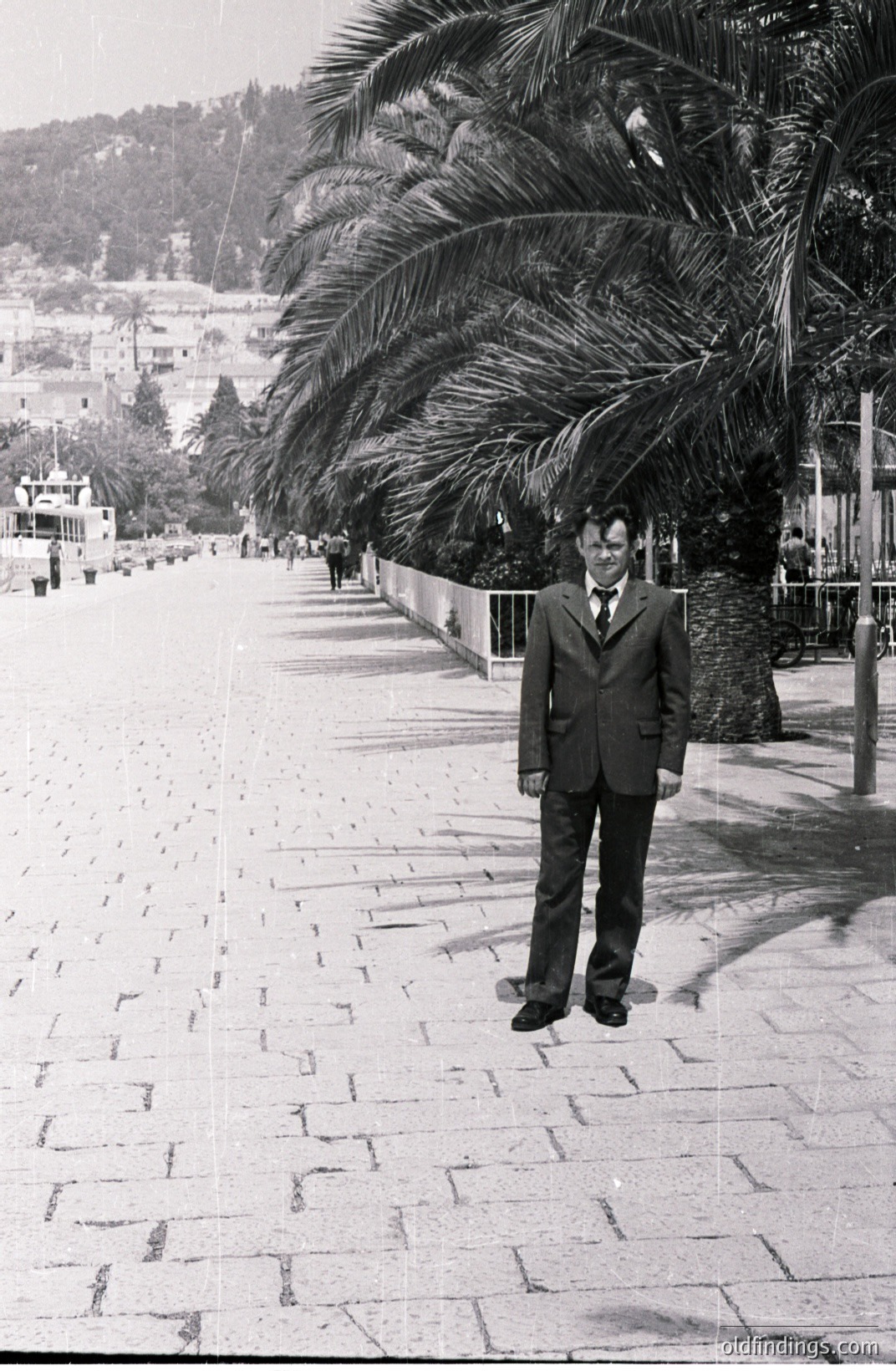 A man in a dark suit and tie stands on a patterned brick pathway, partially obscured by a large palm tree. A coastal town and small boat visible in the distance. Likely a resort area, possibly Mediterranean. Appears to be a candid travel photograph, . Commercial use as a vintage travel design element.