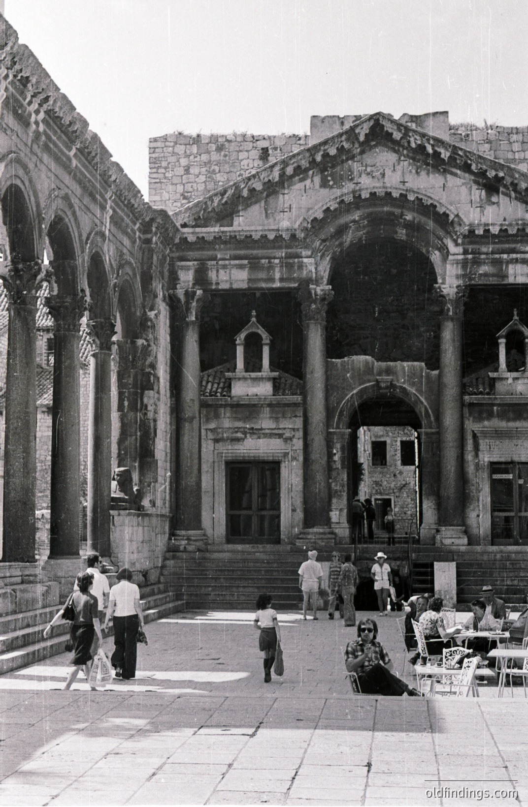 A view of a monumental Roman-era building featuring a large courtyard with arched porticoes and Corinthian columns. People explore the ruins; some seated, others walking. Likely a public space or temple. Appears to be a tourist site.