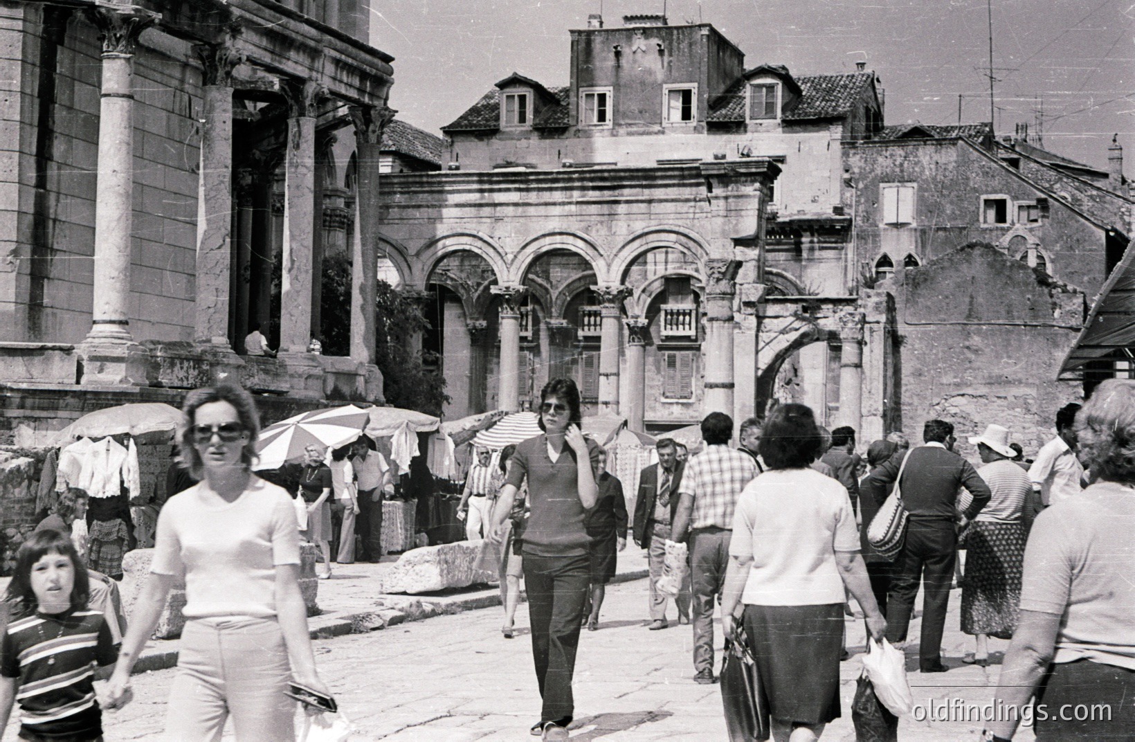 Black and white photo depicting a bustling street scene featuring ancient Roman architecture. Tourists and locals navigate a paved square, overshadowed by a grand archway and remnants of a columned structure. Likely 1970s, possibly in Southern Europe. Captures a moment of daily life against a backdrop of historical significance.