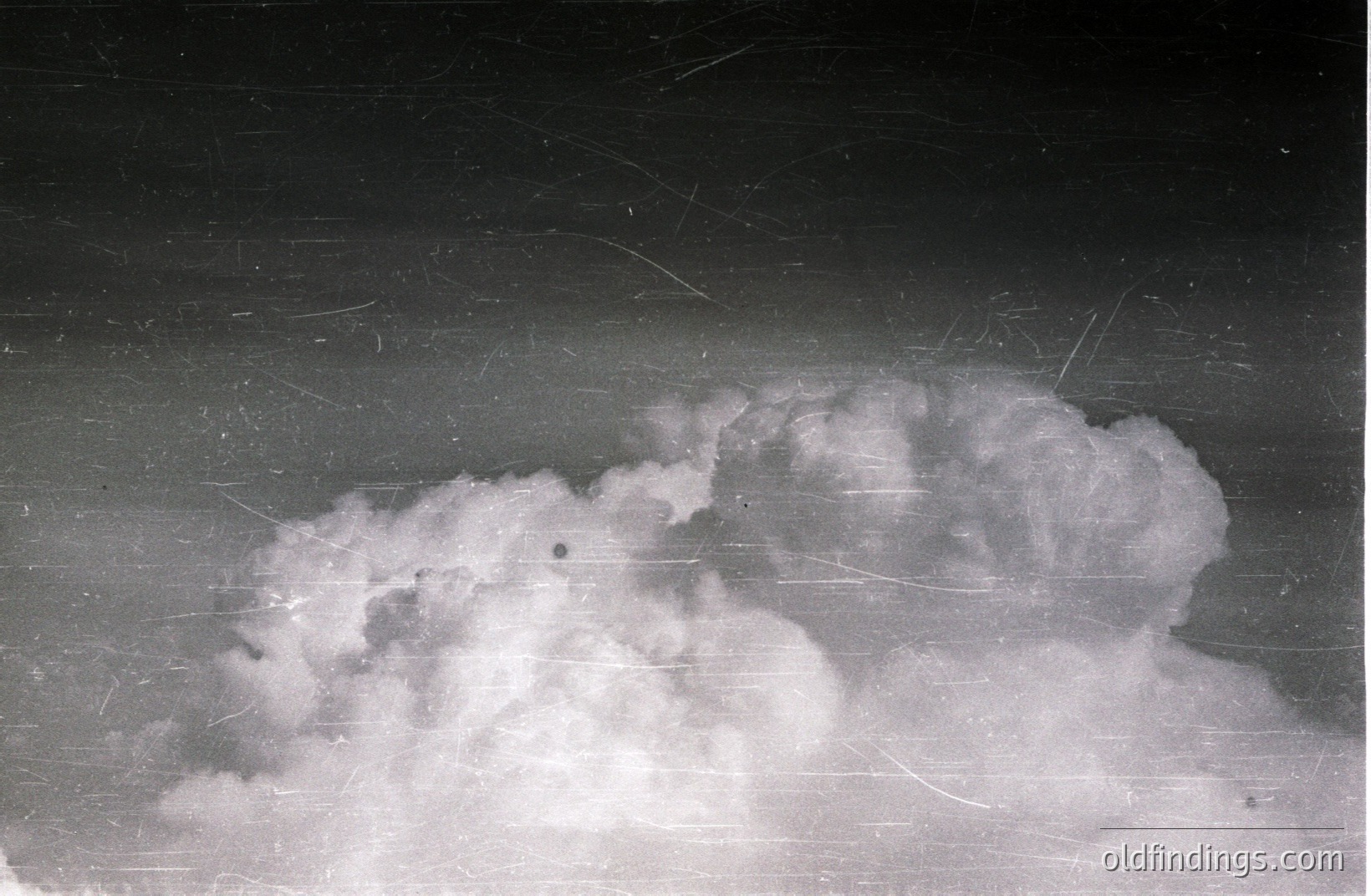 A black and white photograph depicting a dramatic cloud formation, likely a cumulonimbus, dominating the sky. The image exhibits significant age and wear, displaying scratches and dust characteristic of older photographic prints. Strong contrasts & textures. Potential 1940s-1960s era.
