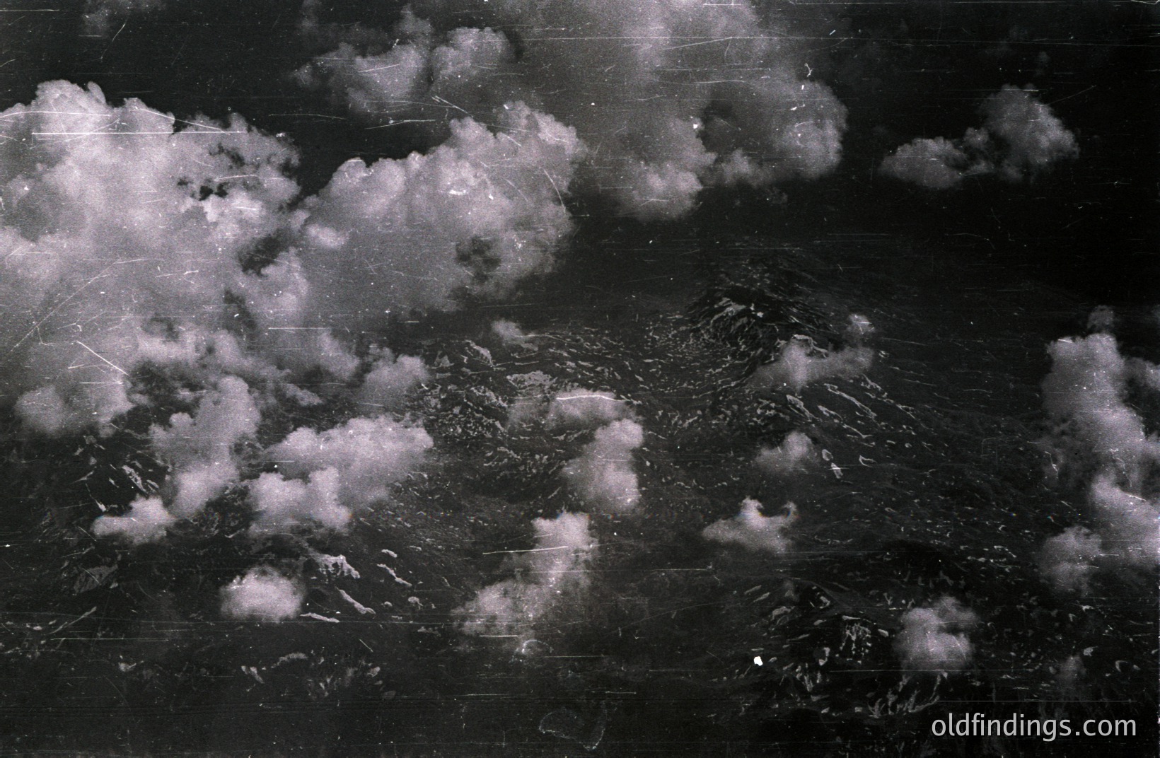 High-altitude aerial view showcases a rugged, mountainous landscape partially obscured by voluminous clouds. Visible textures suggest a rocky terrain. The image exhibits considerable film grain and surface scratches, indicating age. Likely a reconnaissance or survey photograph.