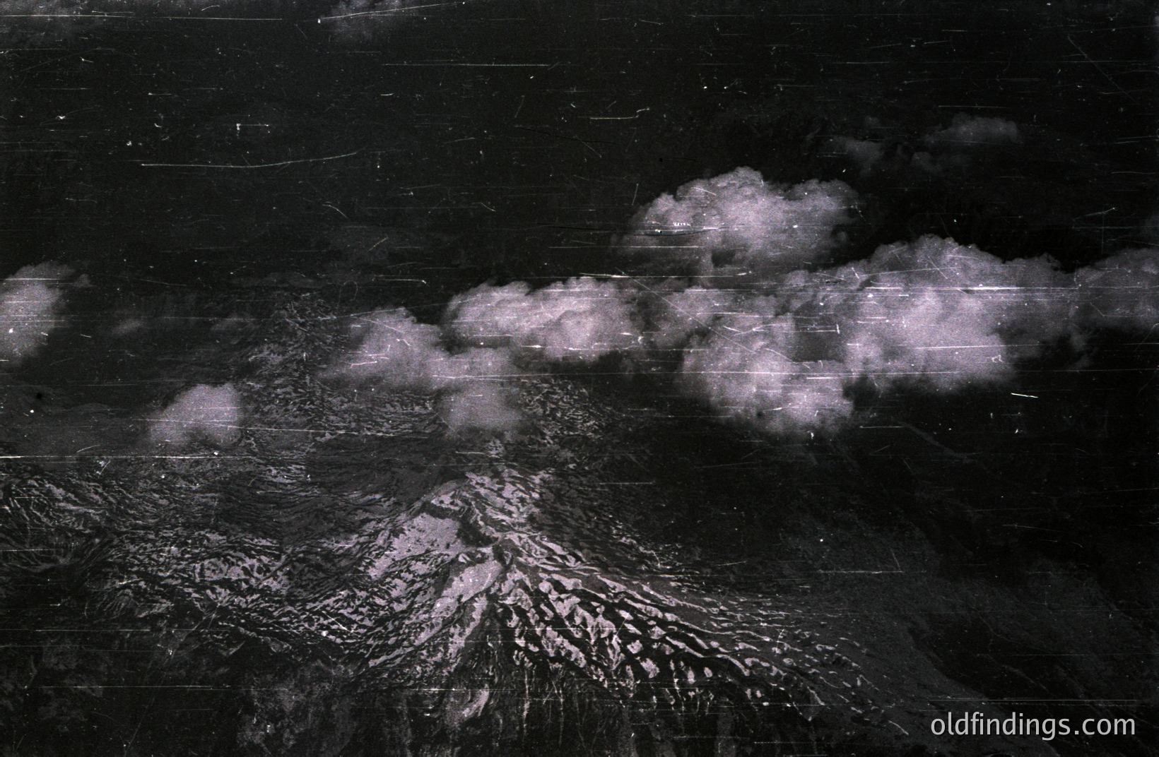 Aerial view of a snow-covered, volcanic peak, partially obscured by swirling cloud formations. The image exhibits significant grain and scratching, consistent with vintage photographic processes. Likely captured during the mid-20th century, possibly for military or scientific documentation. A striking depiction of rugged terrain.