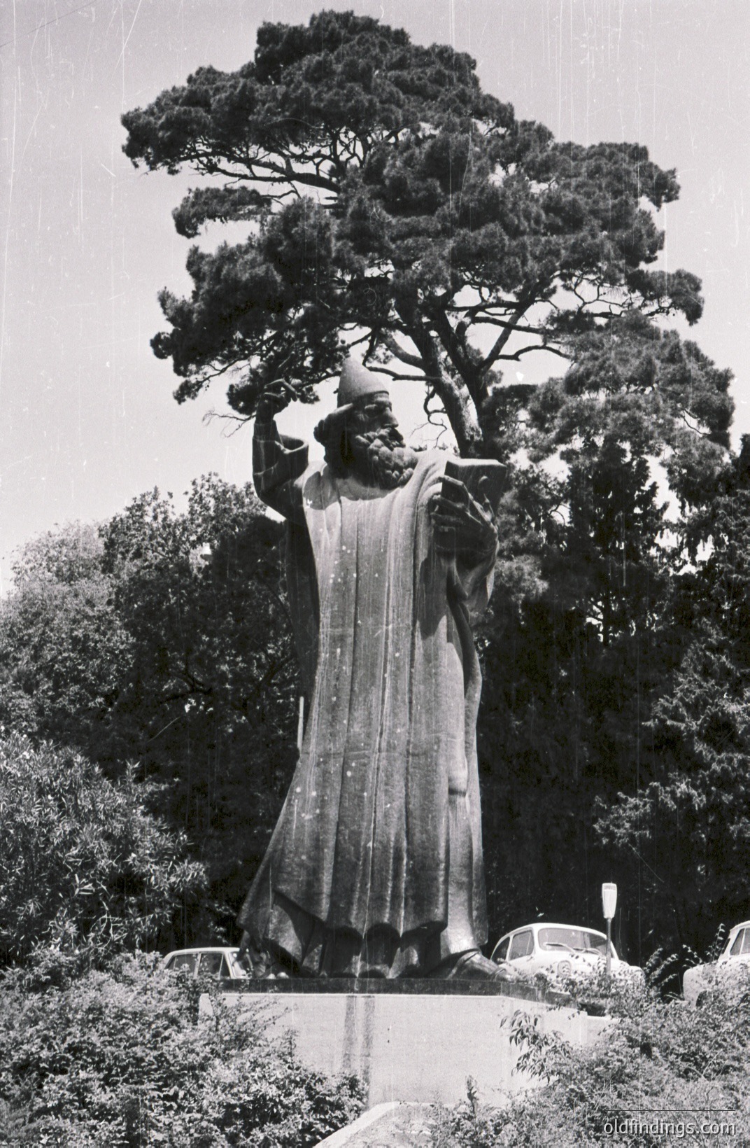 Monumental statue of a robed figure, likely a historical or mythological personage, stands tall against a backdrop of lush greenery. The subject appears to be holding a scroll or book, suggesting a scholar or leader. Likely a public artwork, possibly in a park or plaza. Appears to be a vintage photograph, possibly 1960s-1970s.