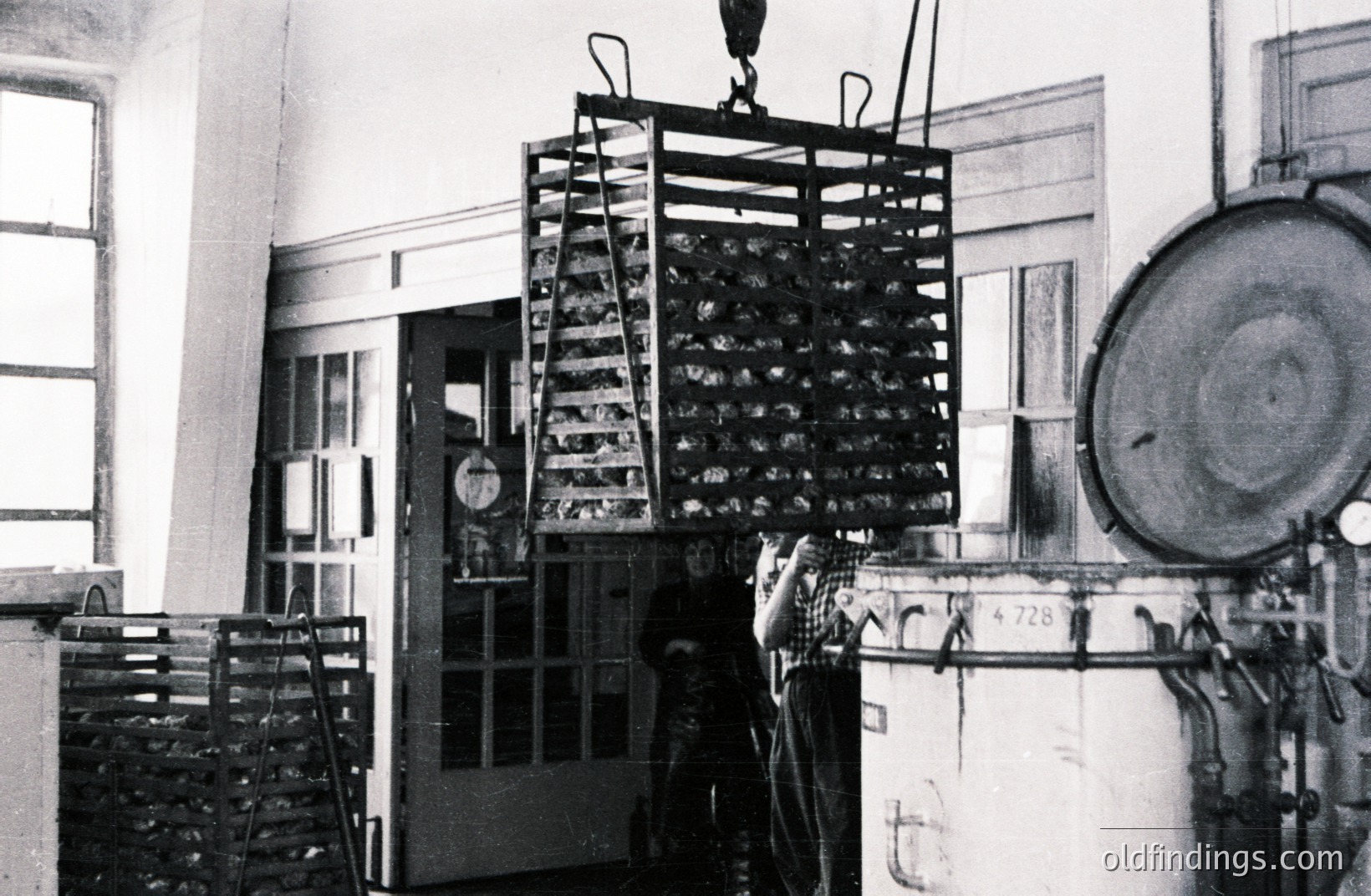 Industrial scene: a large wire basket suspended from a crane mechanism within a factory or processing plant. A boiler sits adjacent, with visible gauges and piping. The room has glass-paned doors and windows. Likely mid-20th century, possibly a canning or food processing facility.