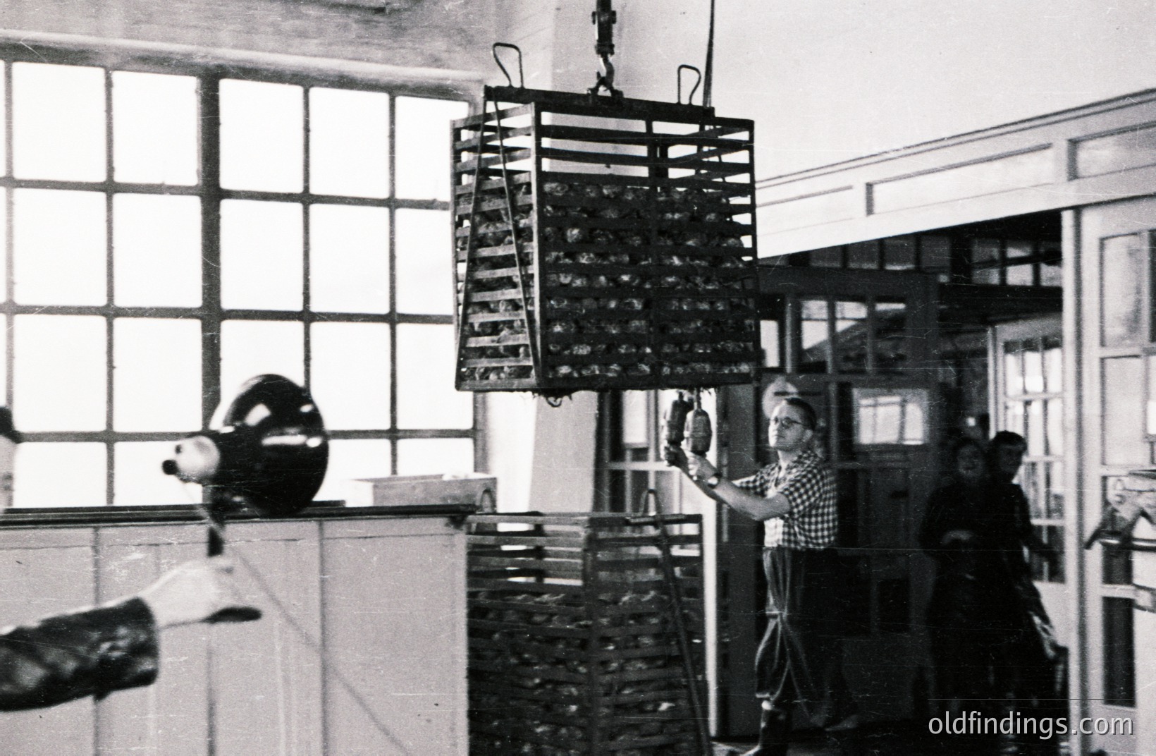 Industrial interior featuring a large, suspended wire rack possibly used for drying or processing. Two workers, one visible full-length, monitor the rack's movement with ropes. Likely a manufacturing setting, possibly food-related. The architecture suggests a mid-20th century industrial space.