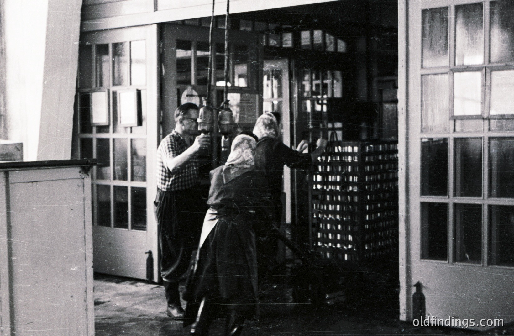 Industrial scene depicting workers near drying racks filled with what appear to be woven textiles. Two men in workwear—one in overalls, the other in a checkered shirt—are positioned beside a tall, windowed structure. The photo's grainy quality suggests a mid-20th century time period. Likely a factory or mill setting.