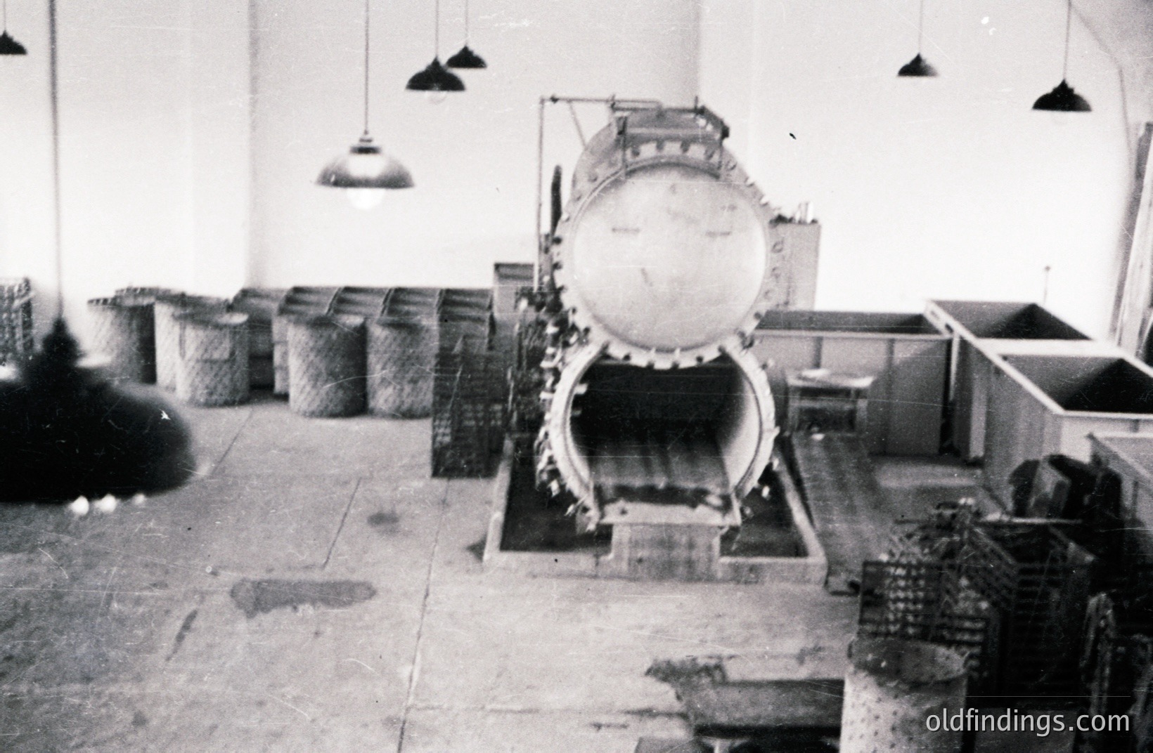 Industrial scene with a large, vintage grain processing machine dominating the space. Woven baskets line the left wall; metal tanks and crates are visible to the right. Low-hanging ceiling lights illuminate the gritty environment. Likely a processing plant, possibly for agricultural products.