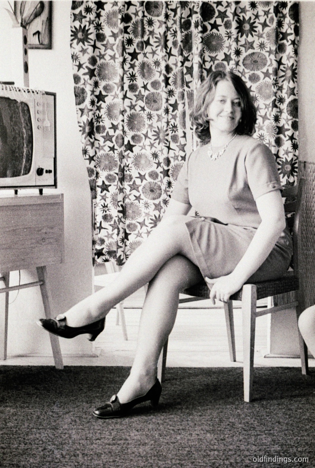 A woman in a mid-length dress and loafers sits poised on a chair, posed in front of floral curtains and a vintage television set. The interior suggests a domestic setting, likely a living room. Style & decor are indicative of the 1960s. A potential stock photo reference for mid-century modern design.