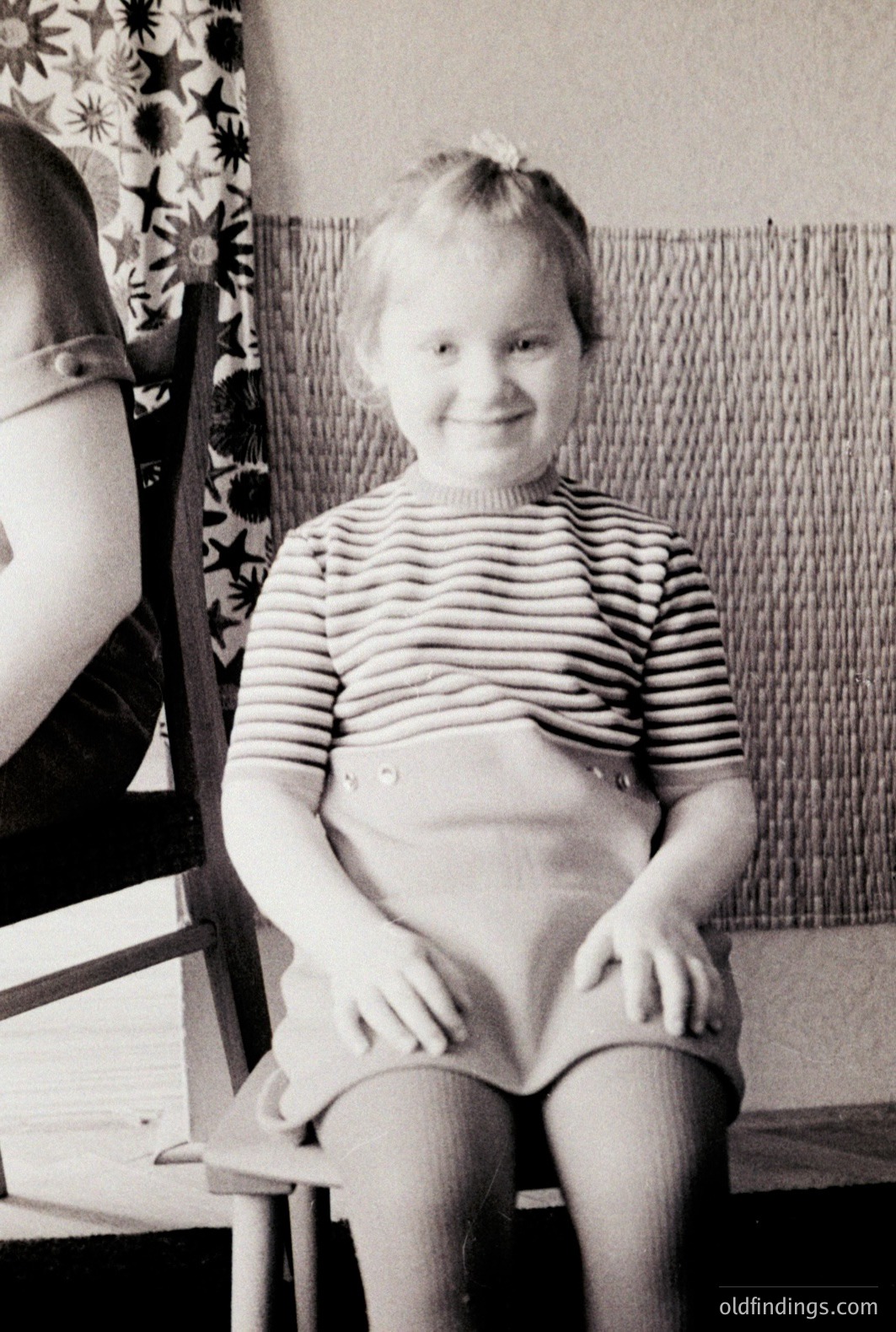 A young girl sits formally in a chair, smiling broadly. She wears a striped top, high-waisted shorts, and a headband. The background includes a woven wall hanging and patterned curtains. Likely a studio or posed portrait, circa 1960s-1970s.