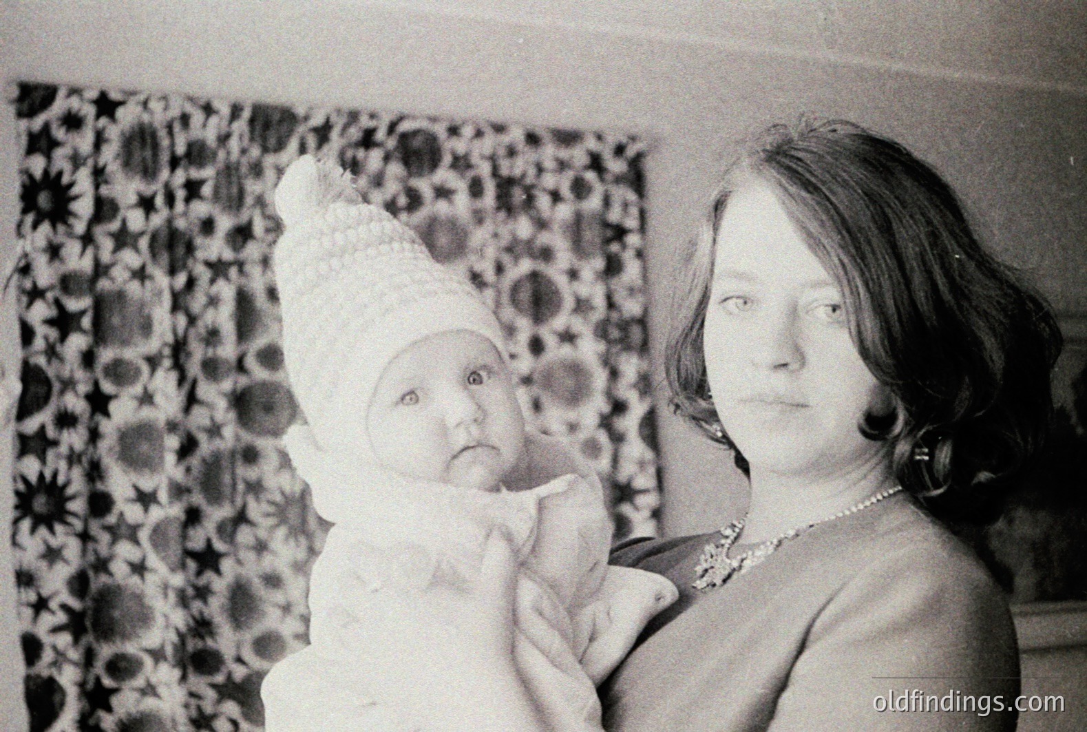 A mother cradles an infant in a vintage portrait. The child wears a fur-trimmed bonnet, while the woman is adorned with a pearl necklace and hoop earrings. Decorative draperies with a floral pattern form the backdrop. Likely a family keepsake from the 1960s or 1970s.