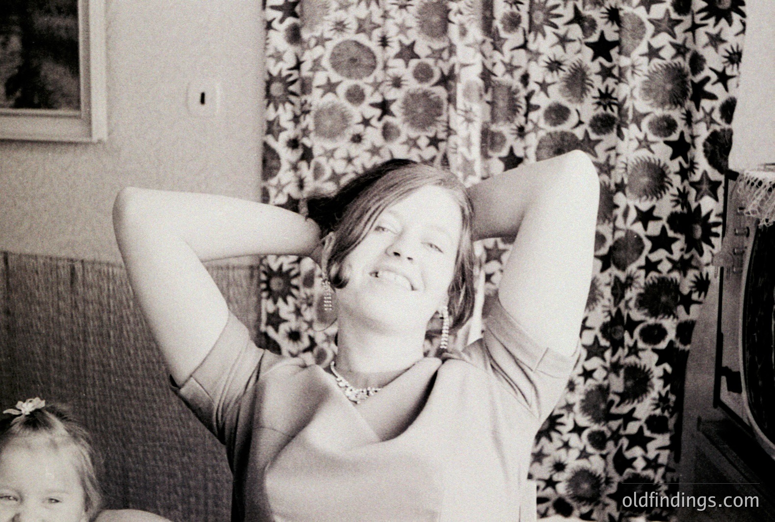 A smiling woman sits on a sofa with her arms behind her head, seemingly relaxed. Detailed floral wallpaper and a vintage television suggest a mid-century interior. A young child’s face is visible in the left foreground. Likely a family snapshot from the 1960s or 70s.