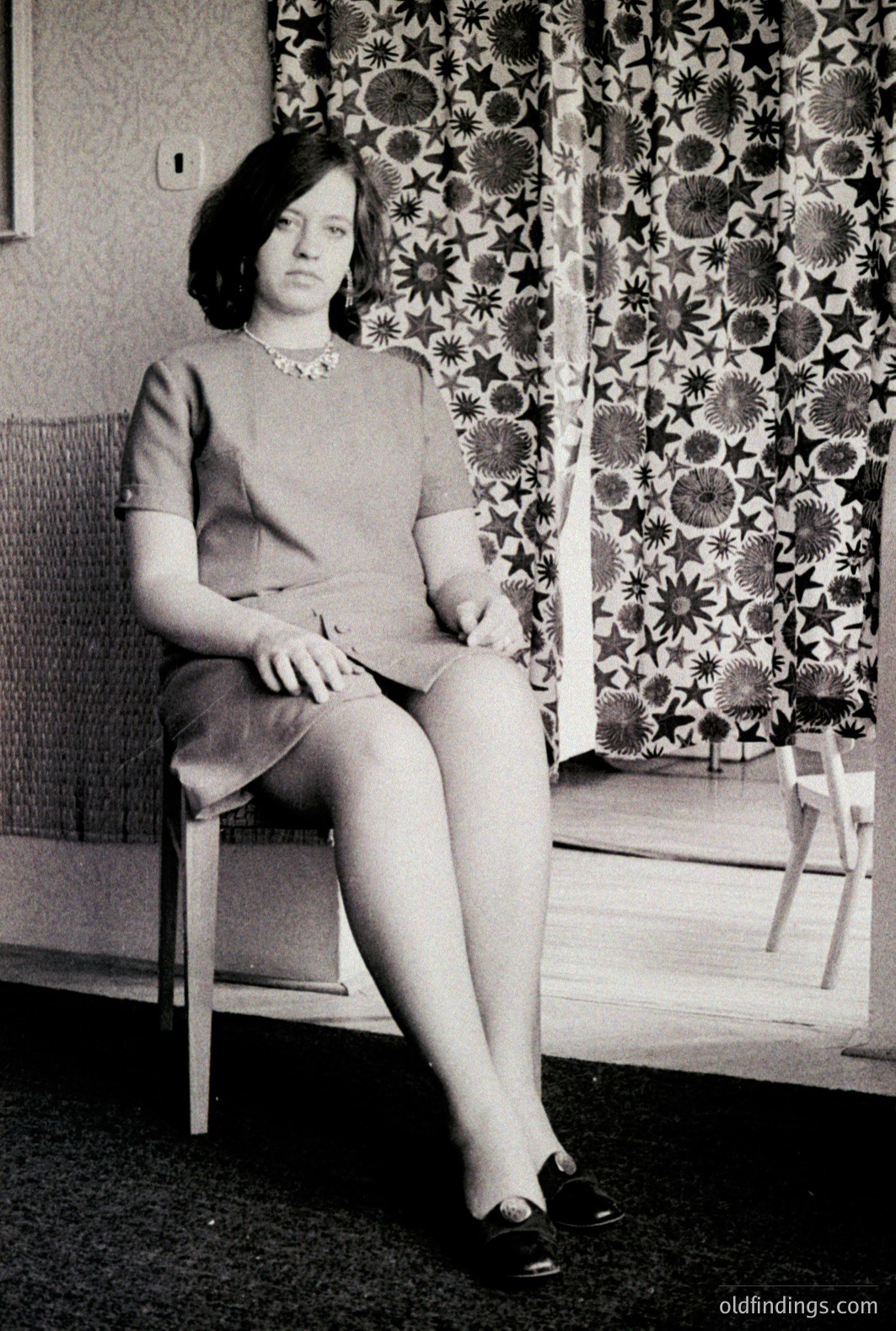 A young woman sits on a chair in a domestic interior. She wears a modest, knee-length dress and a beaded necklace. The background features patterned wallpaper and a textured wall. Likely dating from the 1960s, the image showcases mid-century design aesthetics. Potential for fashion/design reference.