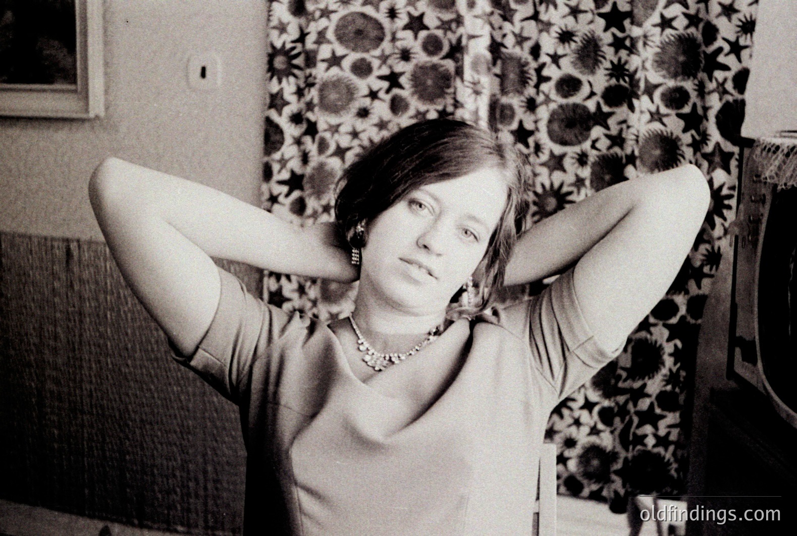 Black and white portrait of a young woman in a V-neck top & statement jewelry, hands behind her head. Likely 1960s/70s fashion aesthetic. Detailed floral wallpaper & simple furniture suggest an interior setting. Potential design reference for vintage style.