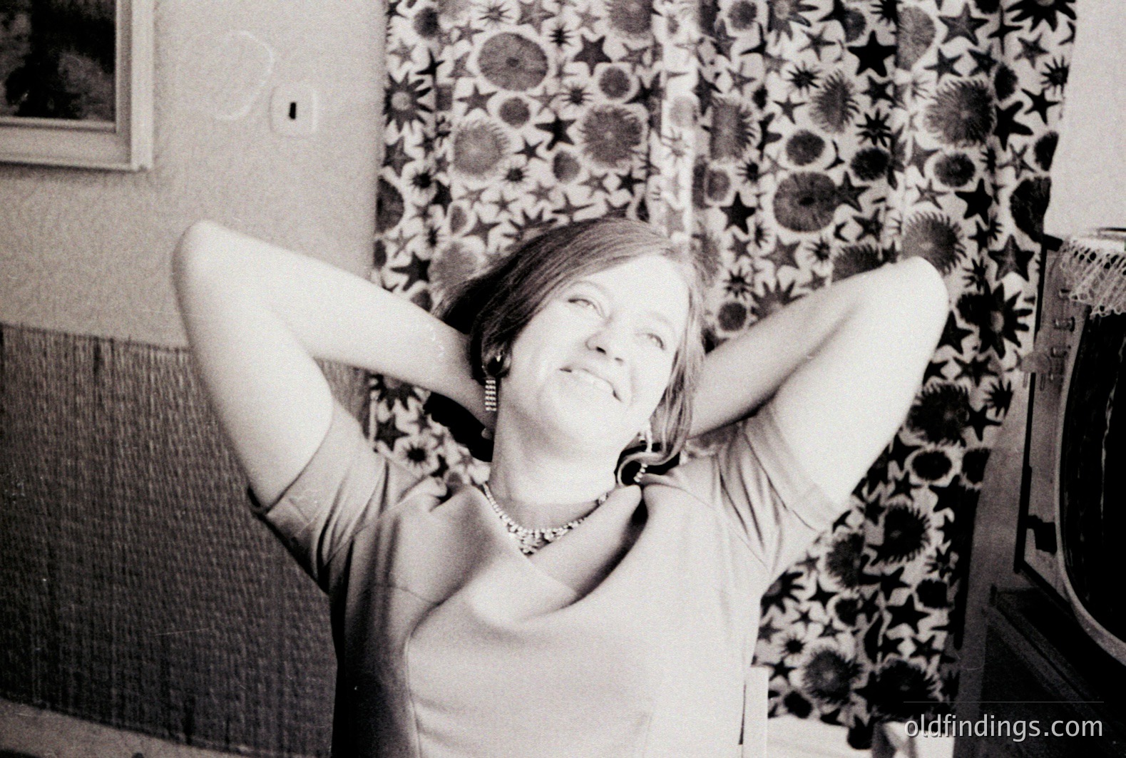A woman with a joyful expression sits, arms raised behind her head. She wears a short-sleeved top and a beaded necklace. The background features a patterned floral wallpaper and a vintage television set. Likely 1970s domestic scene. Potential stock photo for lifestyle or vintage advertising.