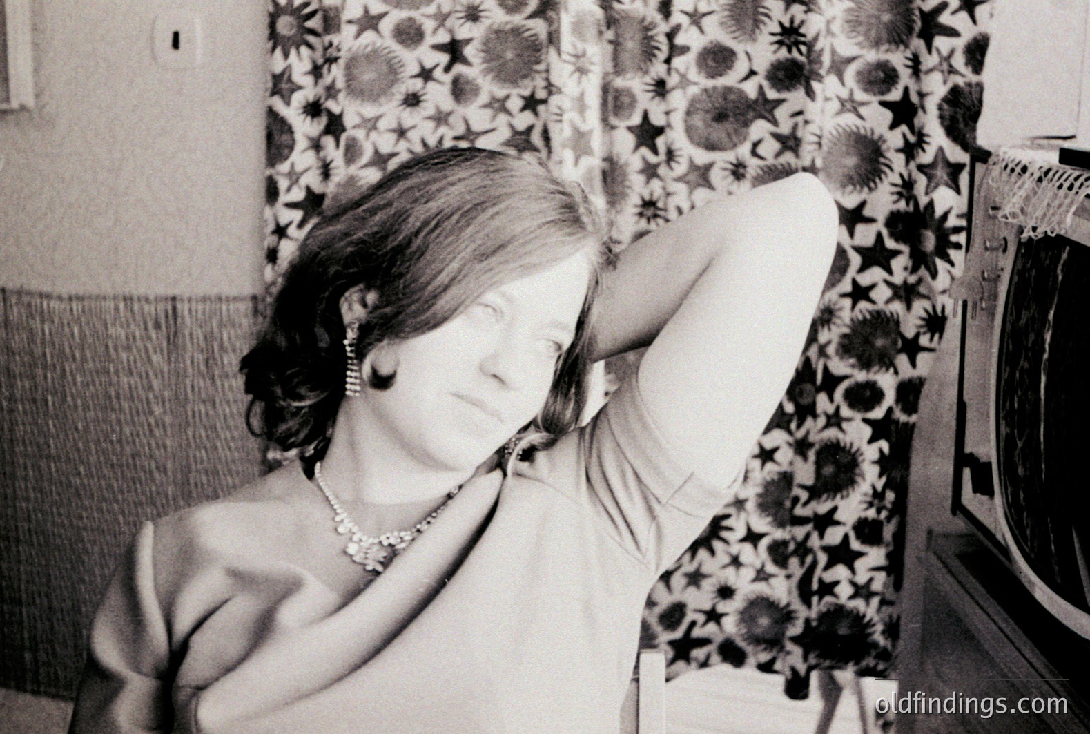 A young woman with vintage styling reclines on a floral-patterned sofa, gesturing towards her armpit. She wears a necklace and earrings. A vintage television set is visible in the background. Likely a domestic interior, 1960s-1970s. Candid portrait with design reference potential.
