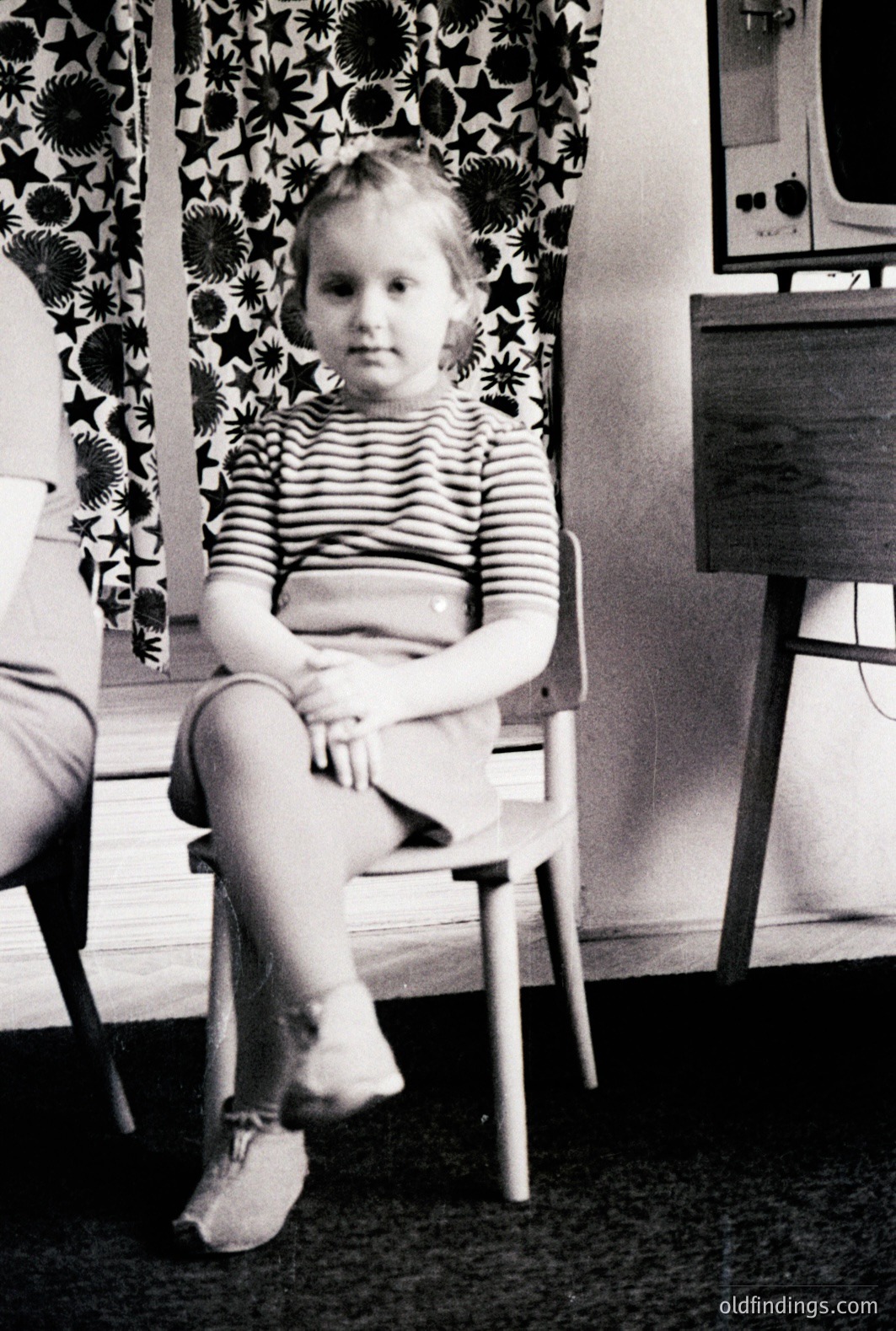 A young girl sits on a wooden chair in a domestic setting. She wears a striped shirt and shorts. A patterned curtain and a mid-century modern radio cabinet are visible. Likely a candid family portrait, c. 1960s-1970s.
