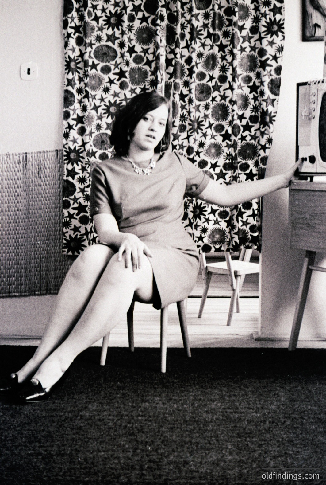 Elegant portrait of a woman in a short-sleeved dress and pearl necklace, seated on a chair with legs extended. The backdrop features a striking floral wallpaper pattern. Likely 1960s interior design; captures mid-century modern aesthetic. Possibly a studio portrait or staged domestic scene.