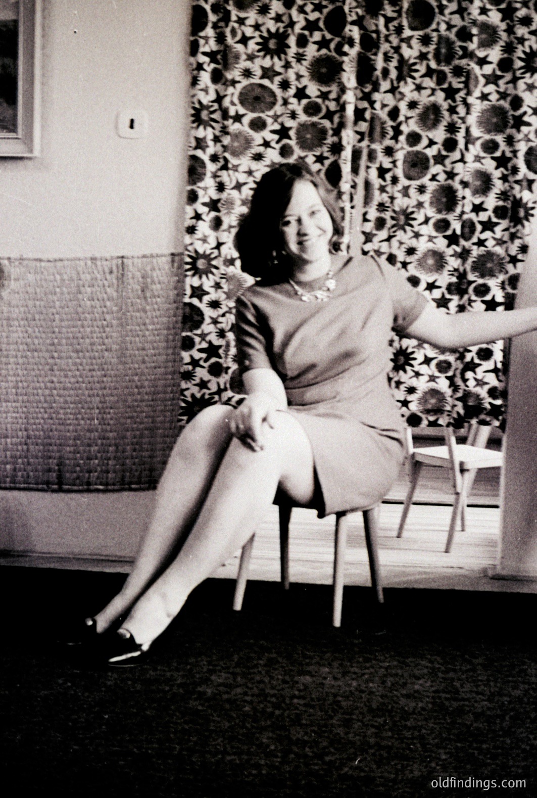 A young woman seated on a chair, posed for a portrait. She wears a short-sleeved dress, stockings, and heels. Behind her, a patterned wallpaper suggests a mid-century interior. Likely 1960s. Captures fashion, home decor, and portrait photography styles of the era.