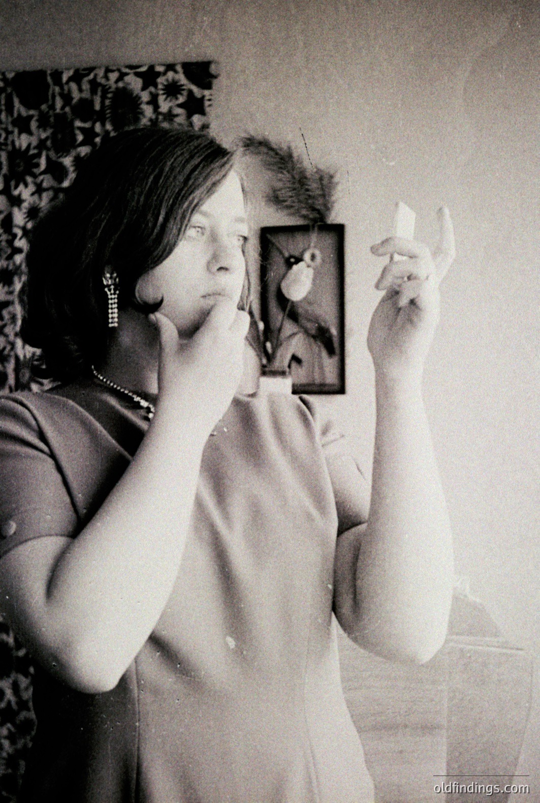 A woman examines her face in a handheld mirror. She wears a sleeveless dress, dangling earrings, and a necklace. A framed botanical print hangs on the wall behind. Likely 1960s style. Classic portrait study.