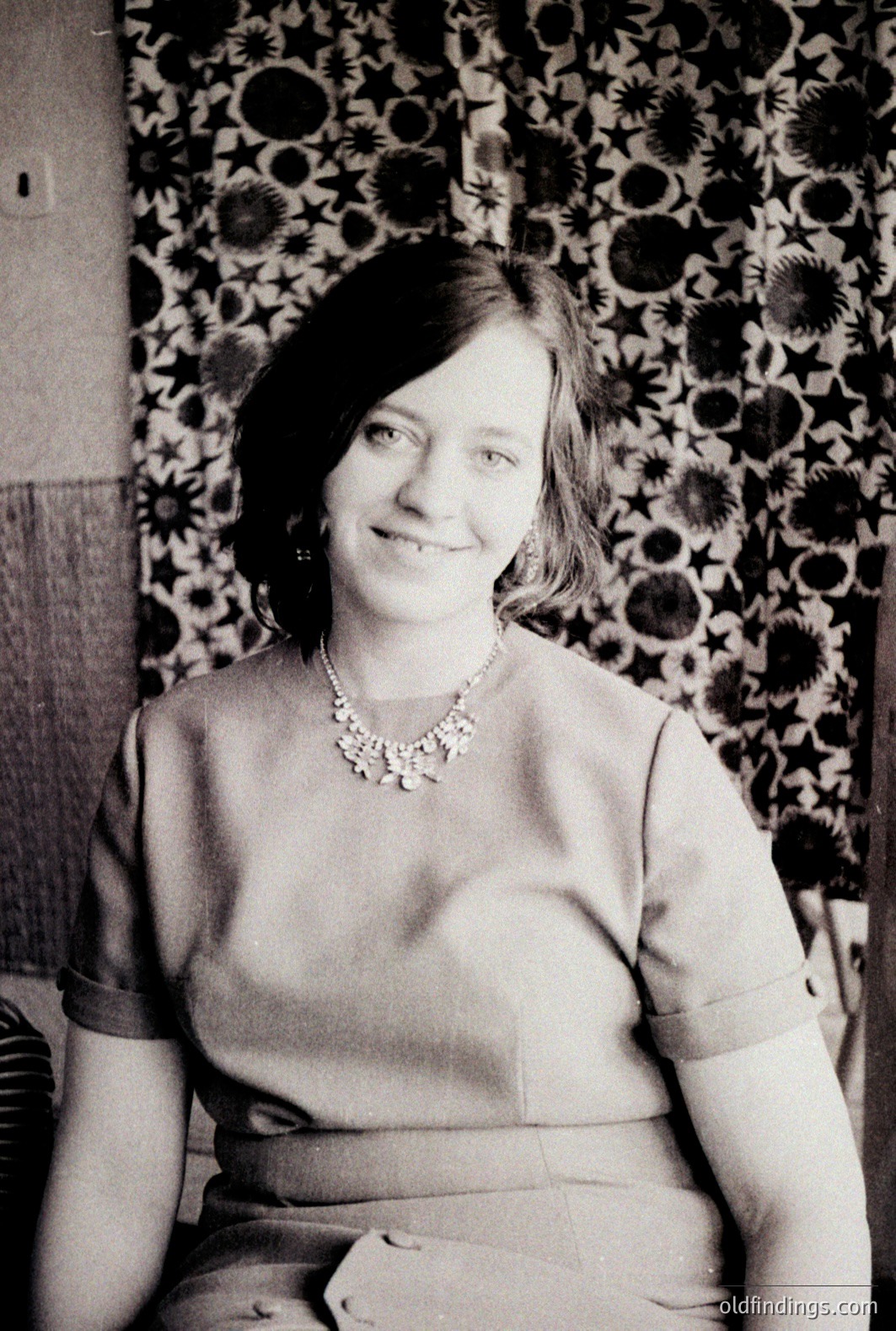 A seated woman smiles directly at the camera. She wears a high-necked, short-sleeved dress and ornate pearl necklace. Bold, circular floral wallpaper forms a patterned background, indicative of a 1960s or 70s interior. Likely a family portrait or informal studio shot.