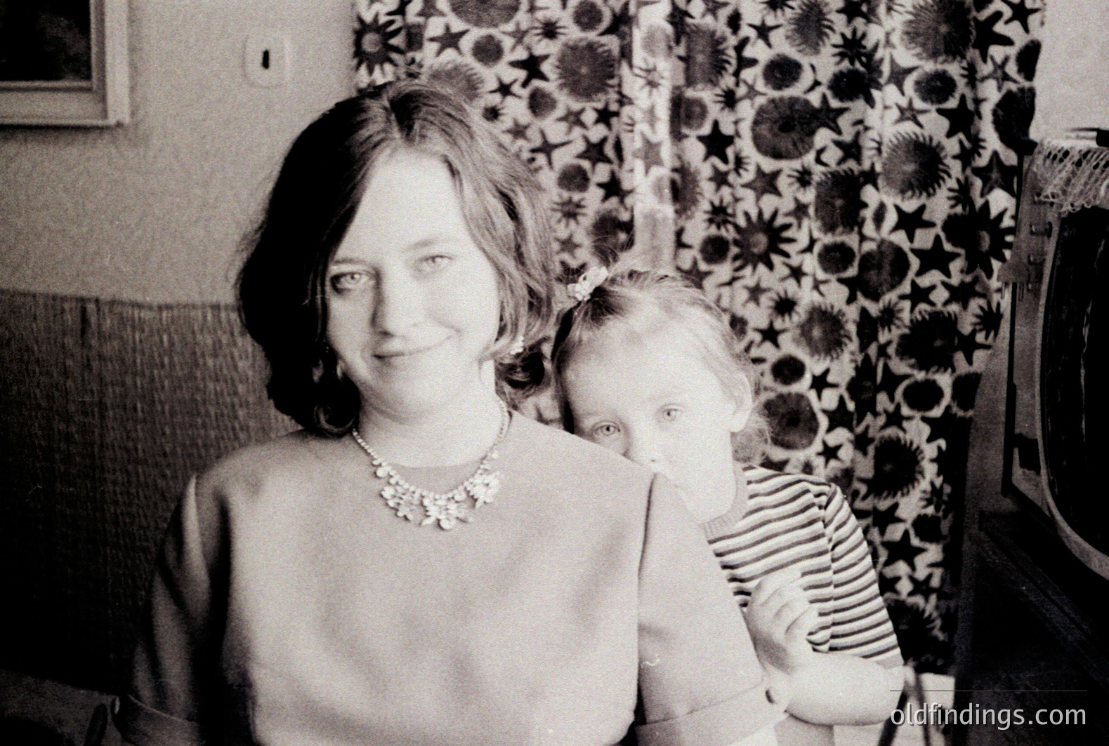 A woman with a pearl necklace poses indoors with a young girl, partially obscured behind her. Likely 1960s/70s, judging by the decor & clothing styles. The patterned wallpaper features dark floral motifs. Appears to be a family snapshot with potential value for genealogy or design reference.