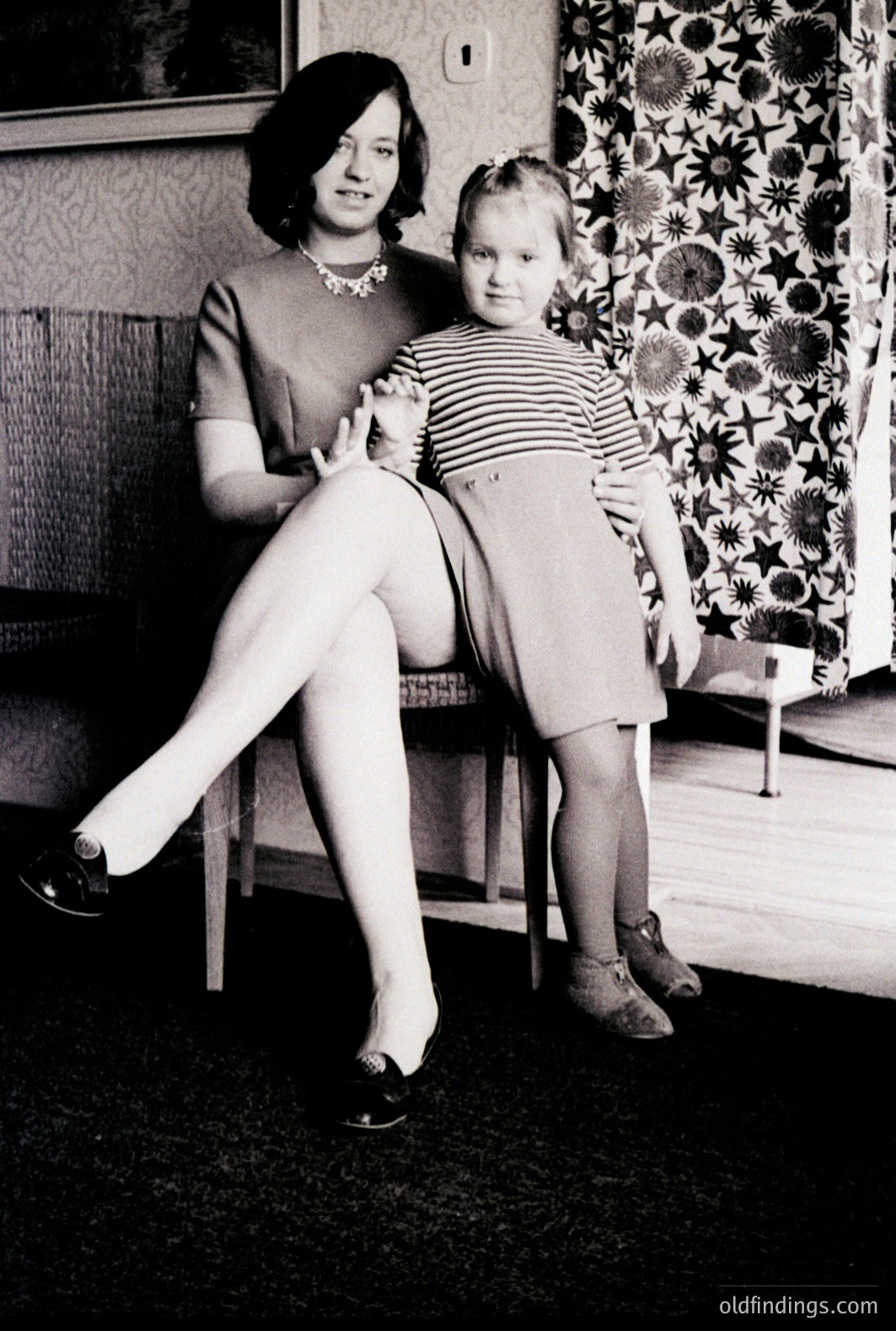 A young woman in a tailored dress & heels sits beside a child wearing a striped dress and slippers. Set in a domestic interior with a patterned wallpaper and dark flooring. Likely a staged portrait, reflecting mid-century family photography.