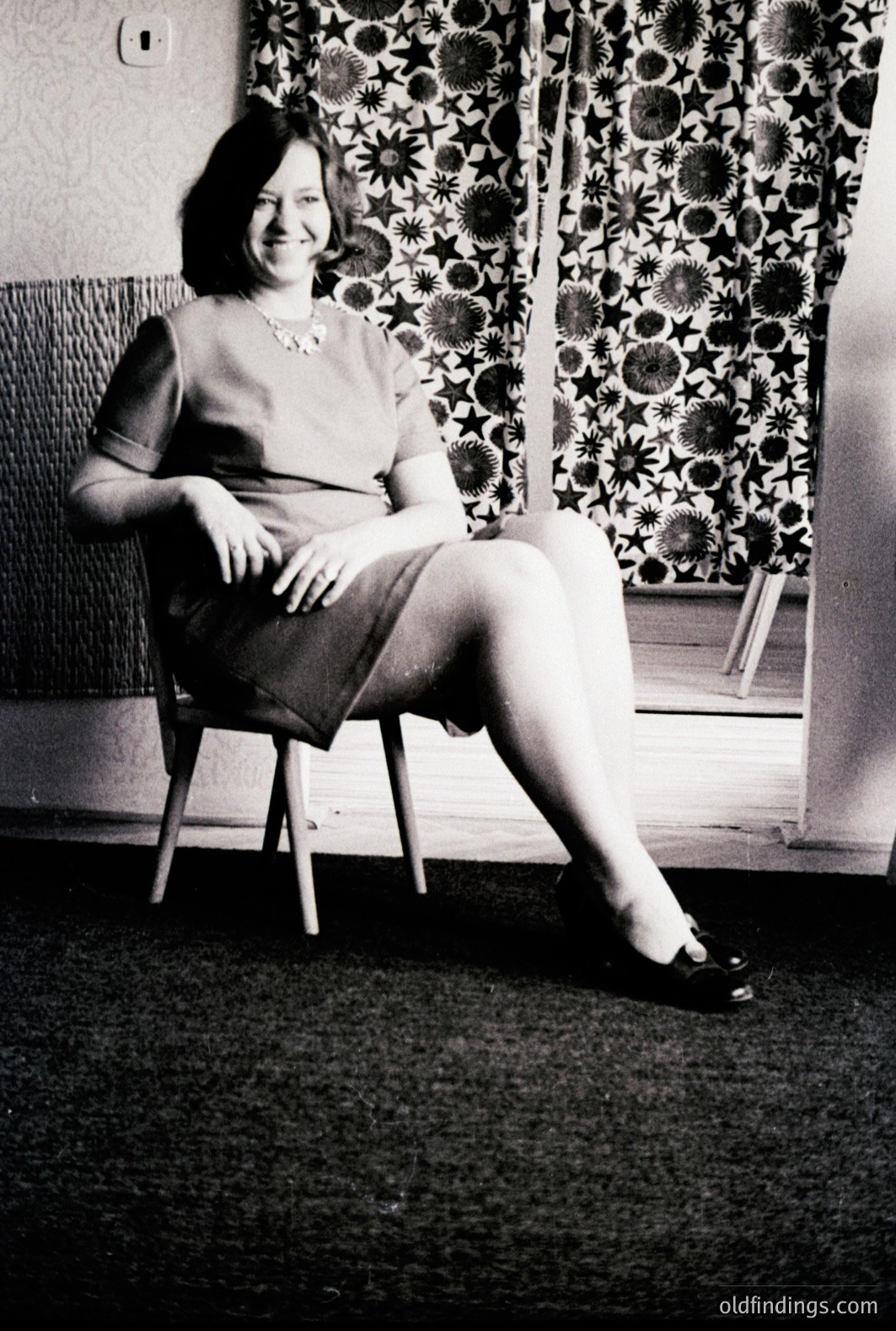 A woman sits in a wooden chair, posed for a studio portrait. She wears a fitted dress, pearl necklace, and dark shoes. Background features patterned wallpaper & a visible light switch. Likely a domestic setting, possibly a studio backdrop. Estimated 1960s-1970s. Commercial value: design reference, family history.
