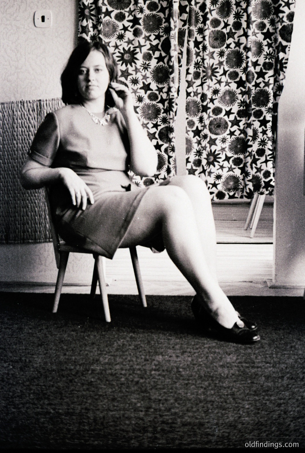 Black and white portrait captures a woman seated in a chair, exhibiting a poised yet contemplative expression. She wears a fitted dress and a pearl necklace, seated on a wooden chair before a patterned curtain backdrop. The decor suggests a domestic interior, circa 1960s-1970s.