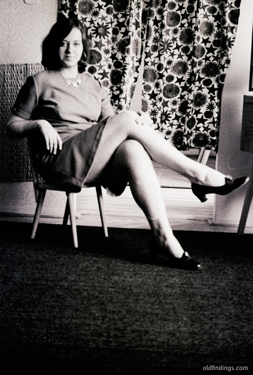 Young woman seated on a simple wooden chair, posed with relaxed posture. She wears a fitted dress and heels. Background features patterned curtains; likely a domestic interior. Style suggests a mid-20th century aesthetic, possibly 1950s-1960s. Strong visual reference for fashion and interior design.