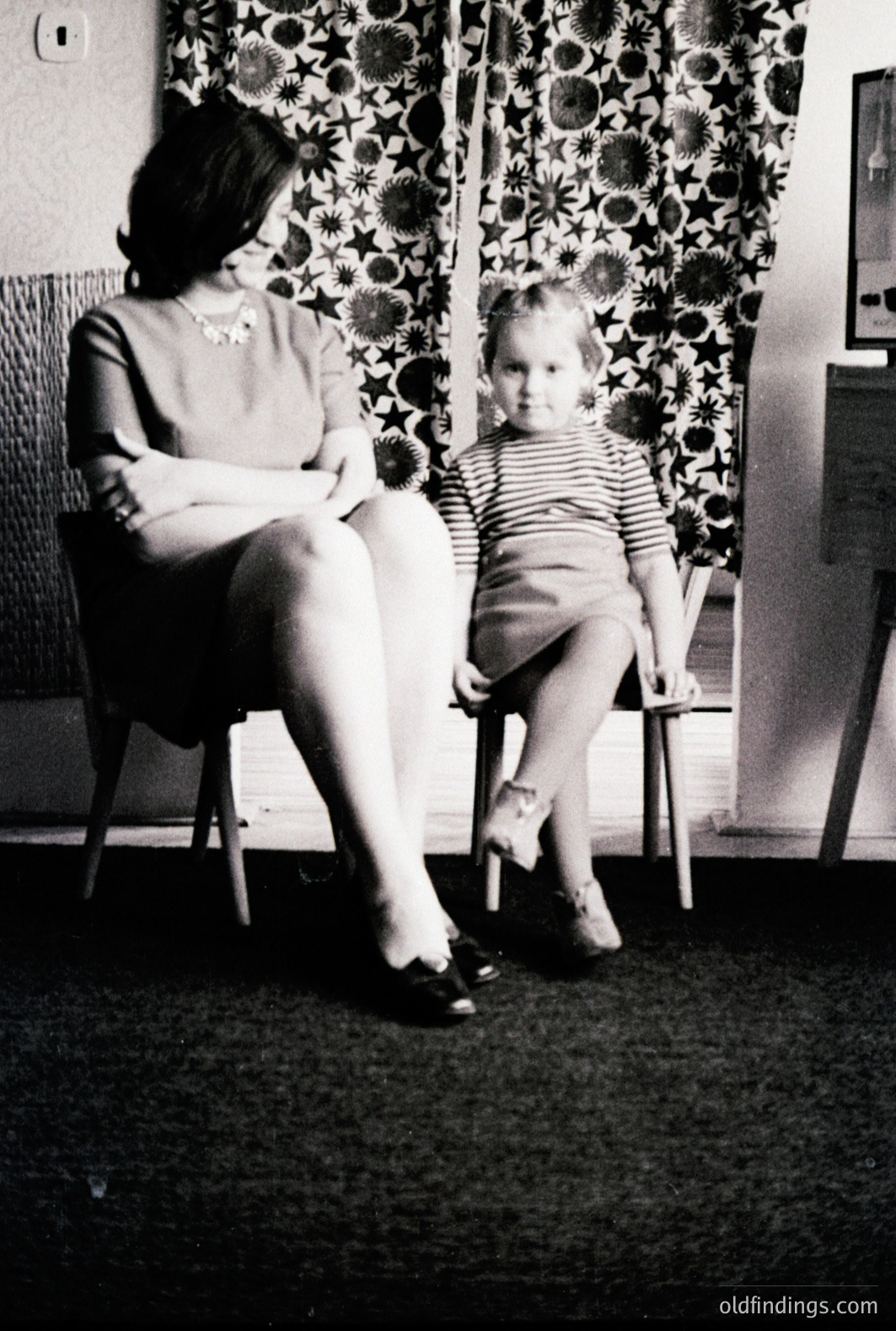 A woman and young child sit on chairs in front of patterned curtains. The woman wears a dress and heels; the child is in a striped shirt and shorts. Likely a mid-century family portrait, capturing a quiet domestic moment. A vintage aesthetic with textural details.