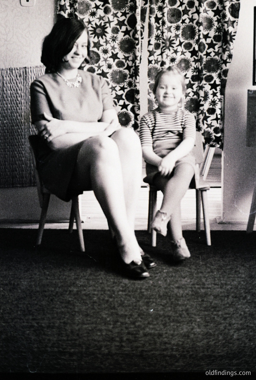 Black and white image featuring a seated woman and child. The woman wears a short-sleeved dress and necklace, the child a striped shirt and tights. Likely 1960s domestic interior, notable for patterned curtains and dark flooring. A simple, intimate moment. Commercial value: design reference, vintage family photography.