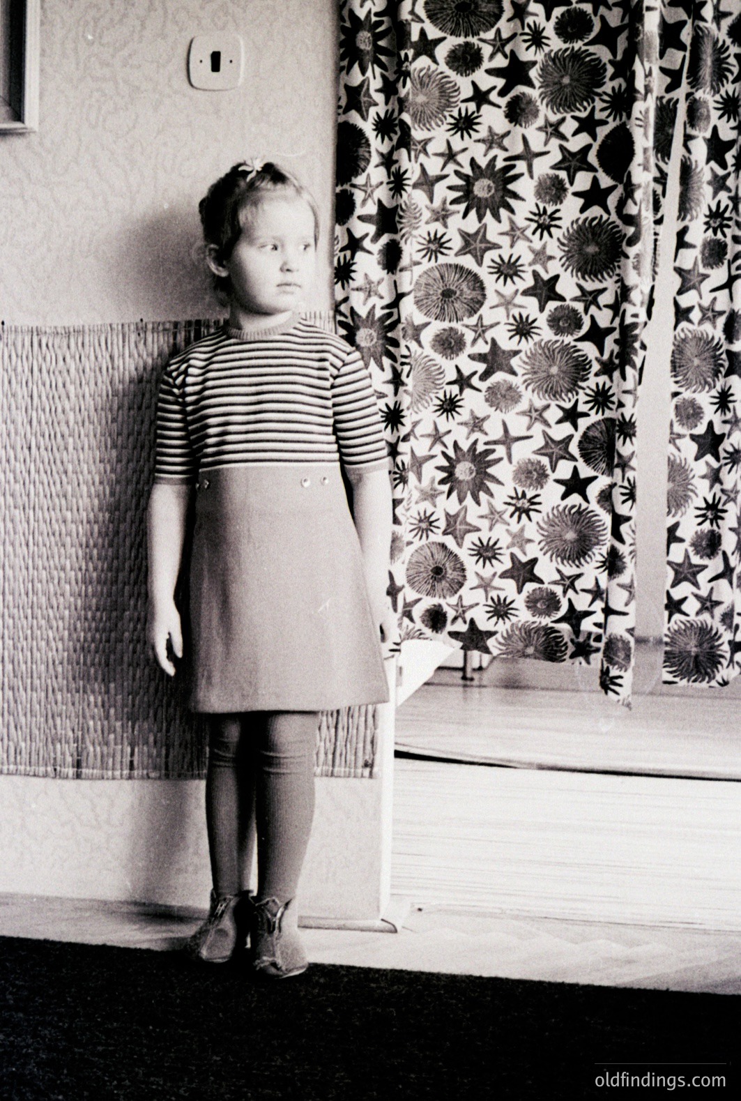 A young girl stands indoors, wearing a striped top and A-line skirt. The backdrop features patterned curtains and textured wall paneling, suggesting a mid-century interior. Minimalist decor and grayscale tones are characteristic of the era. Likely a portrait from the 1960s or 1970s.