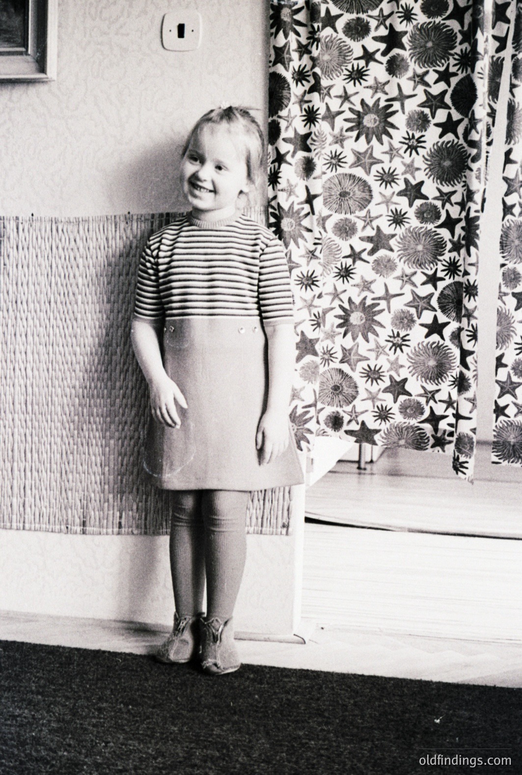 A young girl stands in a domestic interior, wearing a striped top and a simple A-line dress. She smiles directly at the camera. The backdrop features patterned curtains and a textured wall panel. Likely 1960s or 1970s family snapshot. Offers nostalgic charm.