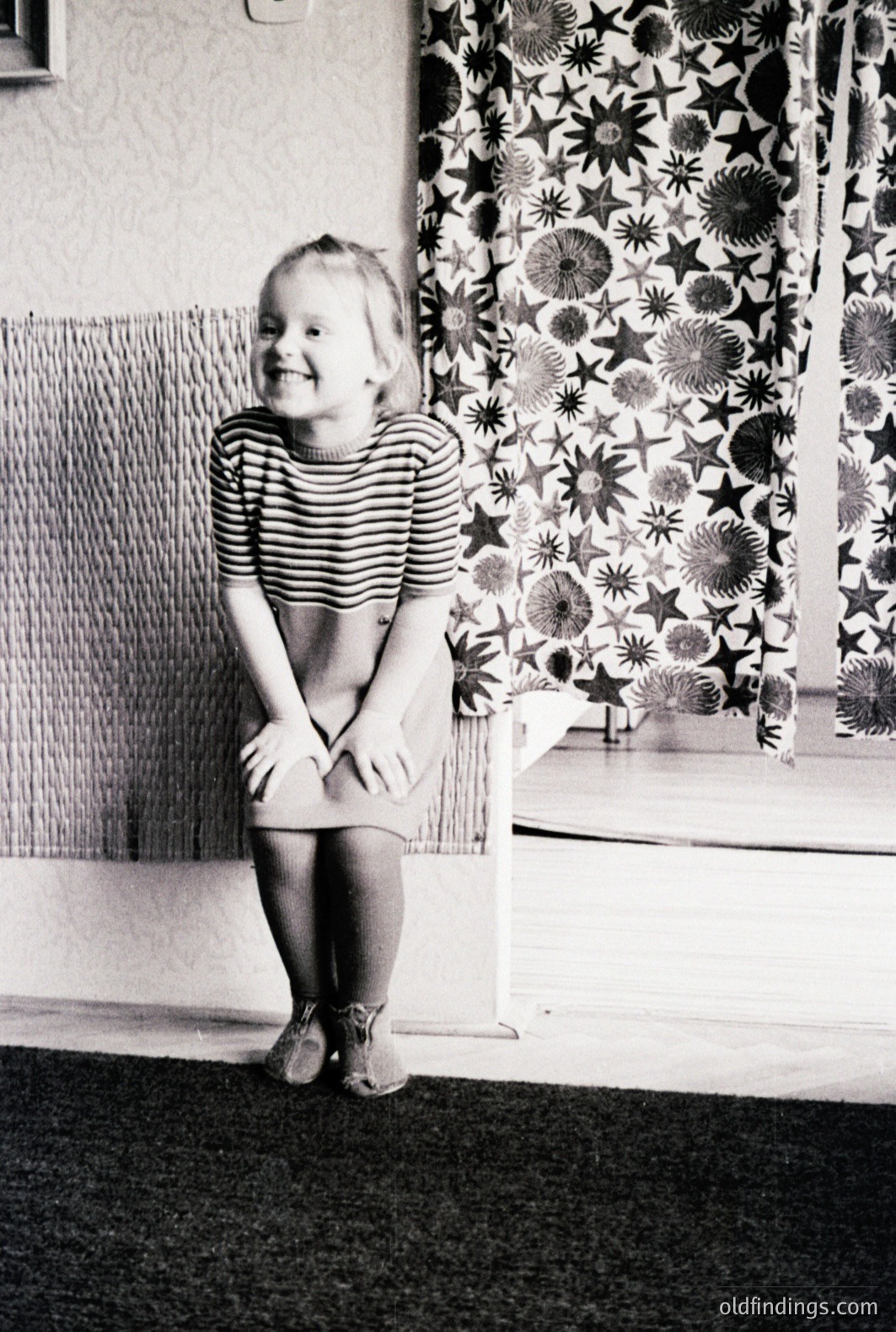 A young girl, likely 5-7 years old, sits on a textured rug, beaming at the camera. She wears a striped top, a dark skirt, and stockings. A vibrant, patterned wallpaper and a chair complete the indoor scene. Likely a family snapshot, 1960s-1970s. Commercial value: vintage childhood, design reference.
