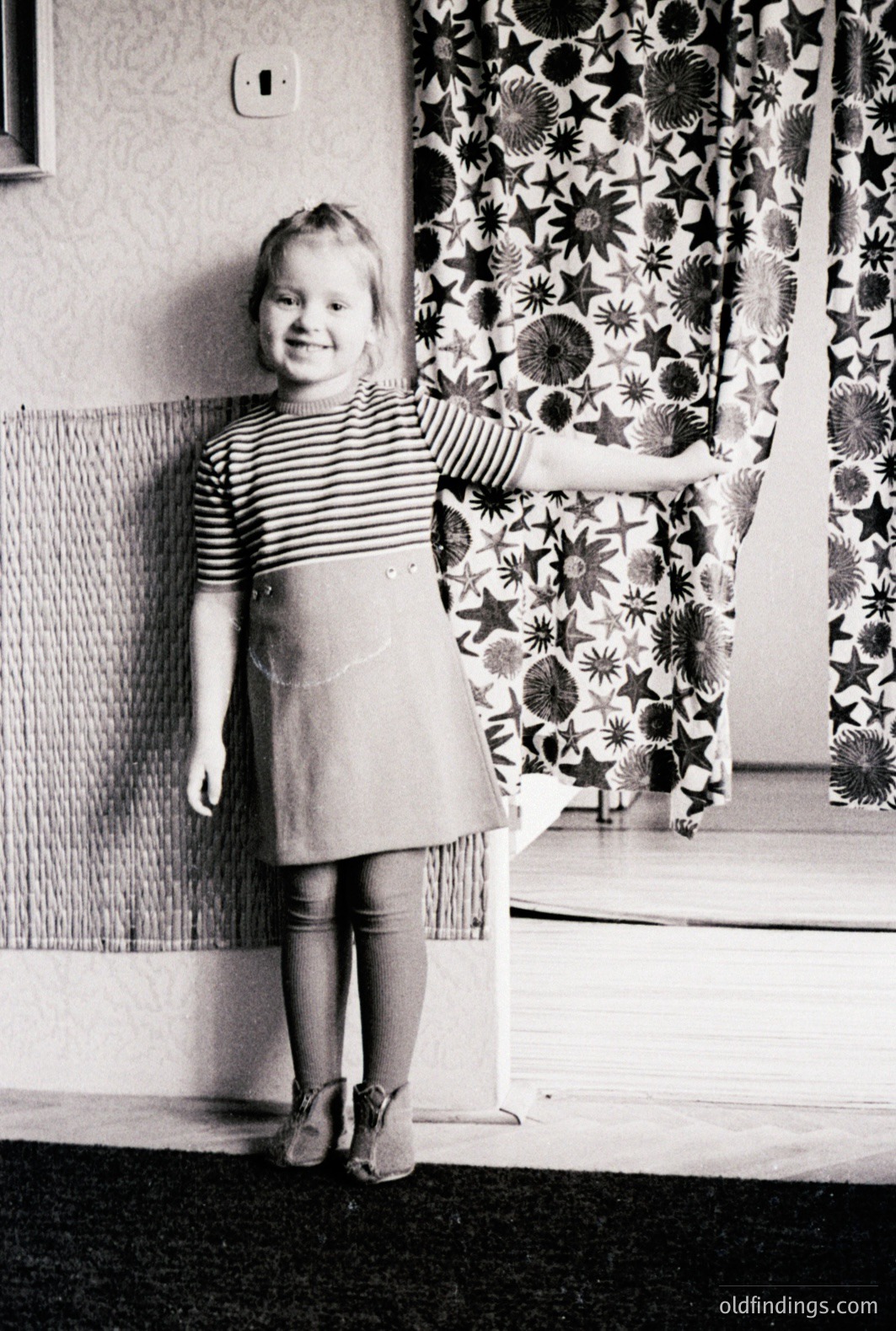 A young girl in a striped shirt and A-line dress stands with a hand on a patterned curtain. Interior scene; likely a home setting. Architectural details suggest mid-century design. Black and white photograph; possibly 1960s or 1970s. Classic domestic scene. Offers strong design reference potential.