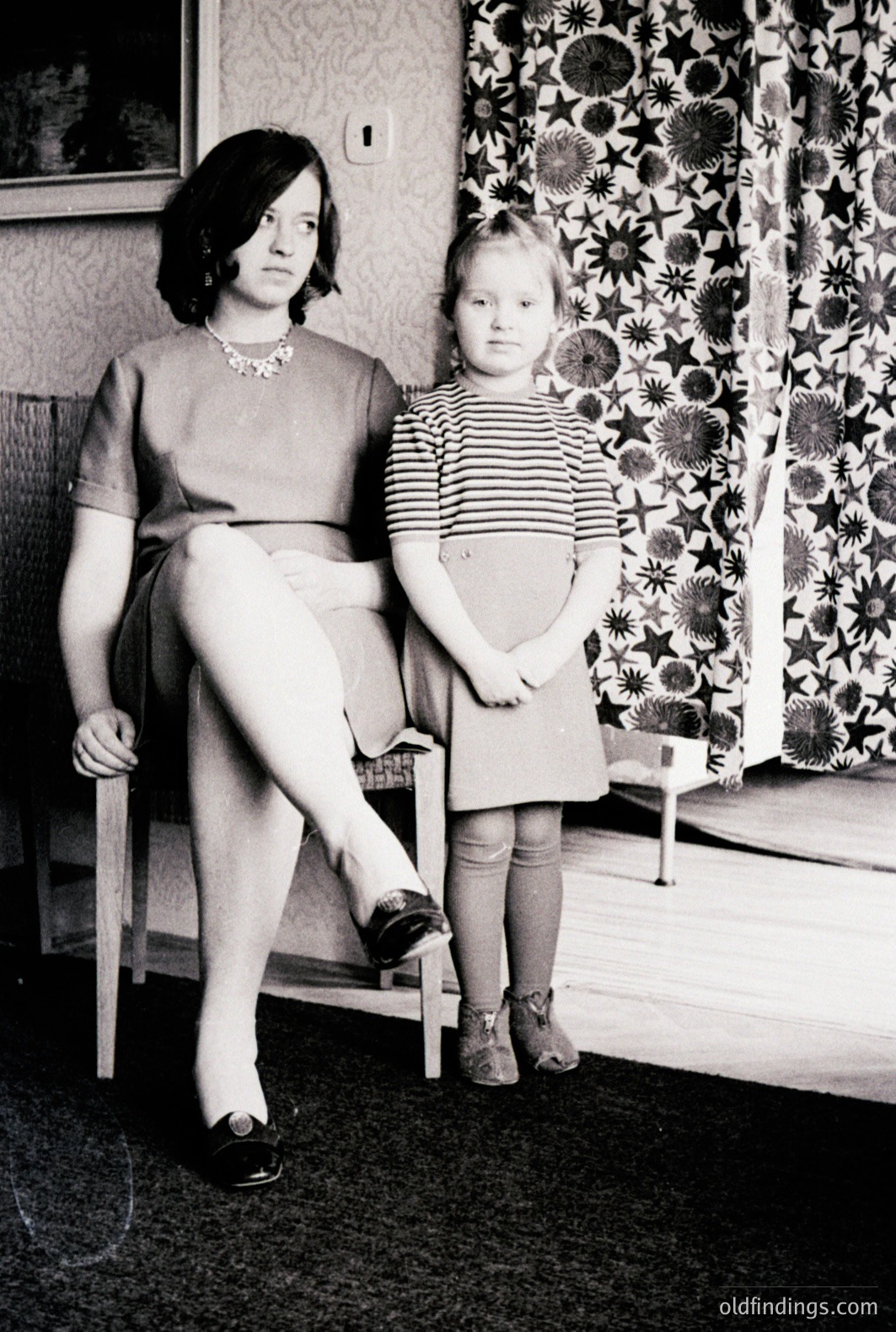Seated woman in a simple dress and patterned tights holds a pose with a young child in a striped dress and matching tights. Interior scene; busy floral wallpaper and visible carpeting. Likely a portrait from the mid-20th century. The woman's jewelry is a notable detail.