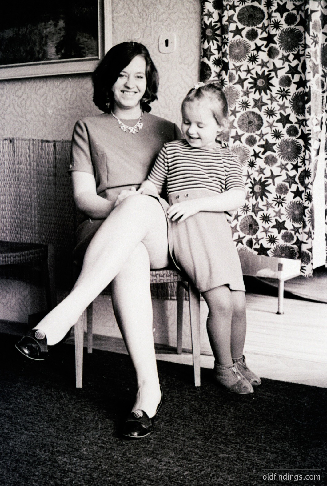 A woman and young girl pose in a home interior. The woman, seated, wears a dark dress and loafers; the girl, in a striped top and matching dress, sits on the woman's lap. A patterned wallpaper and a small side table are visible. Likely a family portrait from the 1960s.