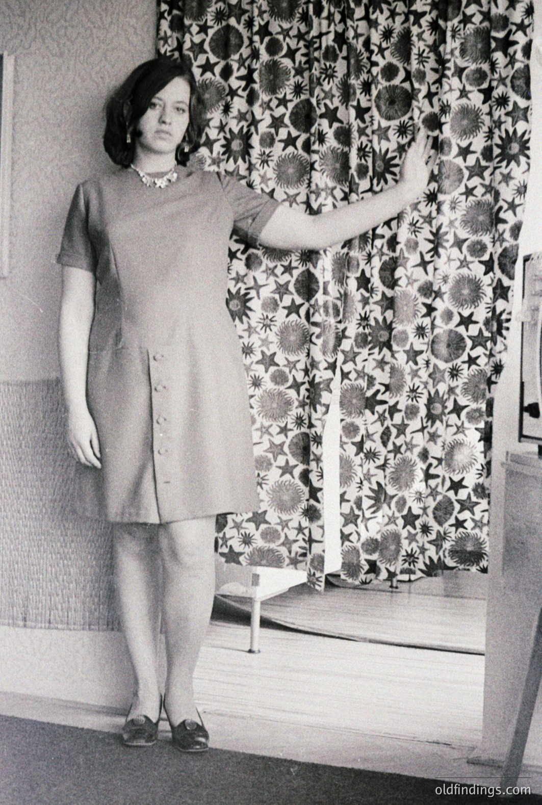 Young woman stands poised in a button-front dress, elevated platform shoes, and pearl necklace. Interior studio portrait with ornate patterned wallpaper. Likely a fashion photograph, circa 1960s or 1970s. Studio equipment visible in background indicates professional setting.