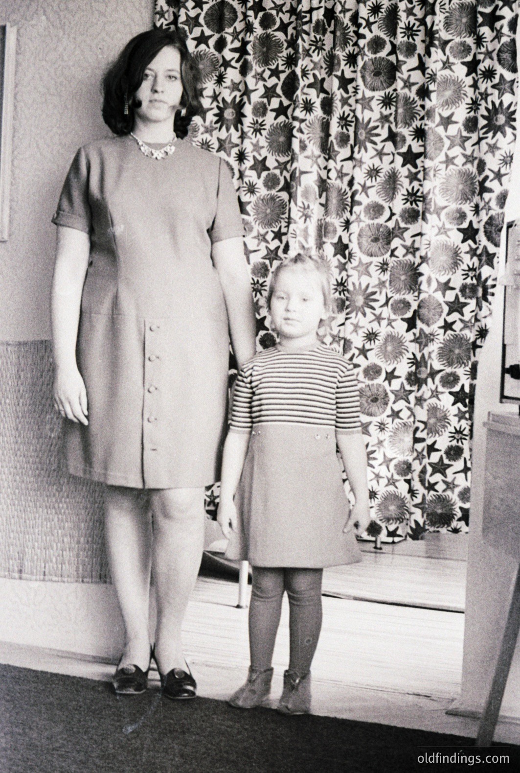 A young woman and child stand before a patterned wall. The woman wears a buttoned dress with a shallow neckline, accessorized with a beaded necklace. The child wears a striped top and a matching skirt with buttons. Interior, likely 1960s, simple composition. Strong potential for design reference.