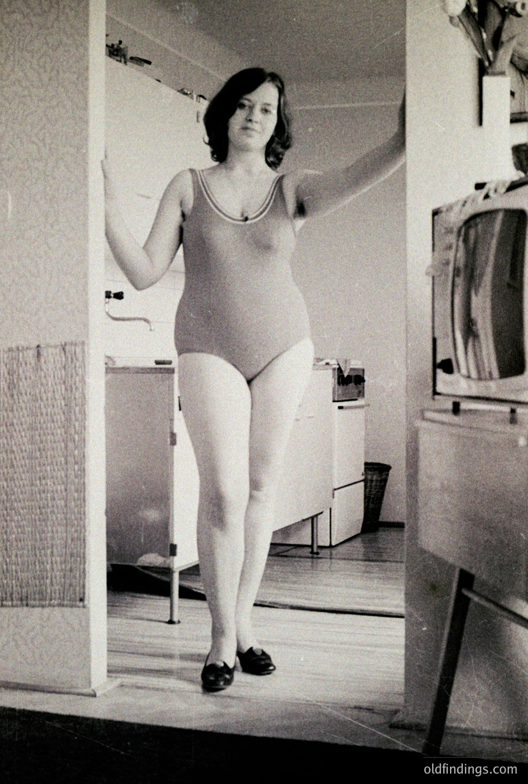 A woman in a one-piece swimsuit poses in what appears to be a bathroom or changing room. Textured wallpaper and tiled flooring are visible. Likely a candid snapshot from the 1960s, the image exhibits characteristics of amateur photography from the era. Commercial potential for fashion/style reference.