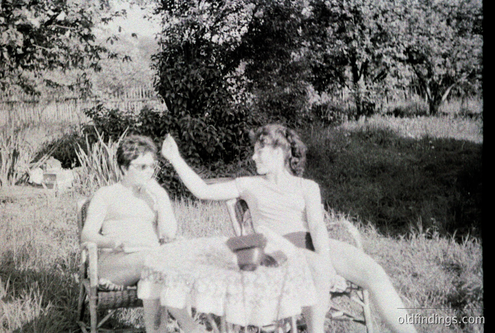 Two young people relax in wicker chairs, casually posed on a grassy lawn with lush foliage in the background. The individual on the right extends a hand, while the person on the left holds an object near their chest. Appears to be a candid, mid-century snapshot.