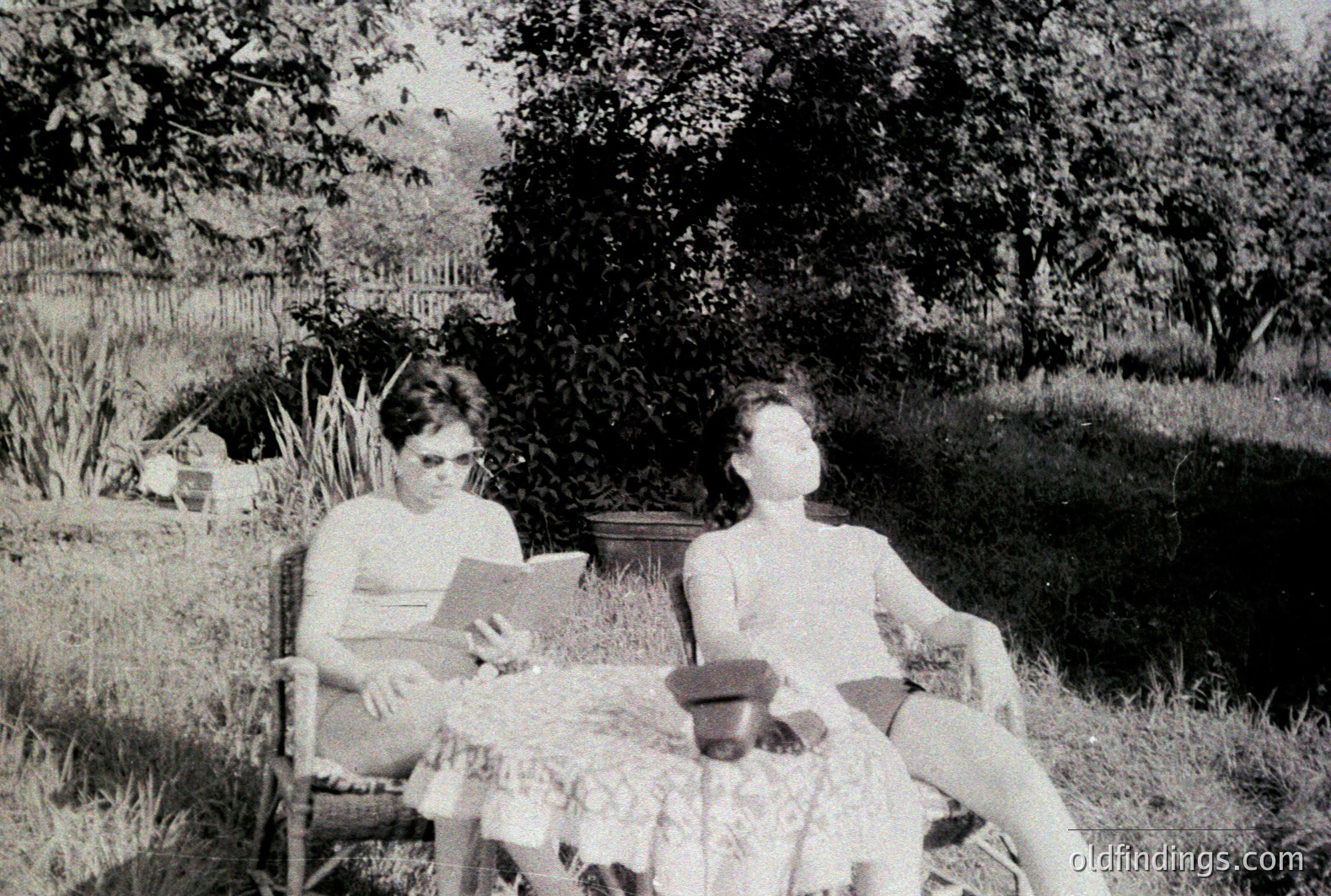 Two figures relax outdoors in a garden setting. One, seated and reading, wears a short-sleeved shirt and dark shorts. The other reclines with a hat nearby. Lush vegetation and a wooden fence form the backdrop. Likely a family snapshot, circa 1950s-1970s.