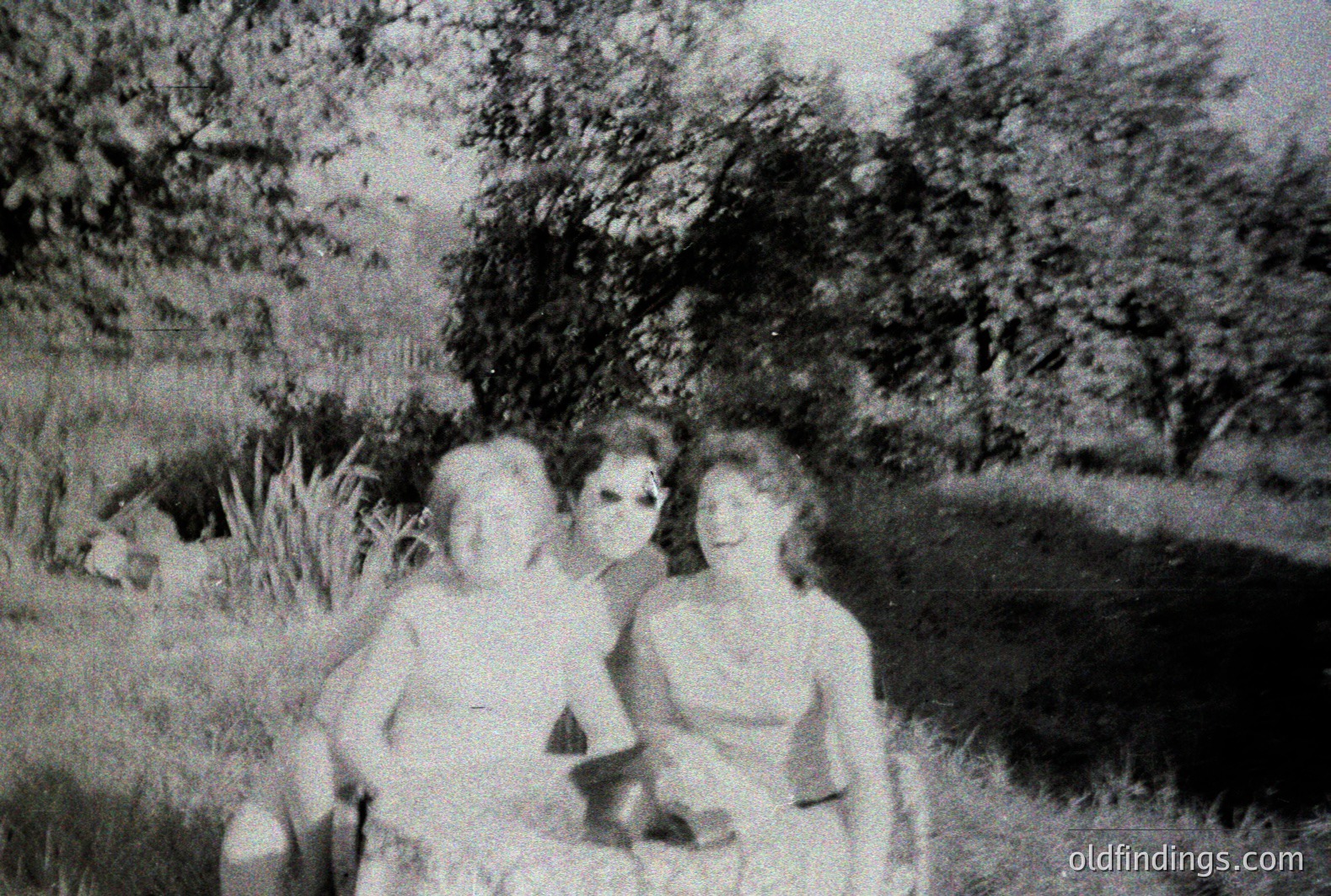 Two women seated on a small hill with tall grass and dense foliage in the background. Both appear to be wearing swimwear. Appears to be a snapshot, possibly a vacation photo from the 1950s or 60s. Grainy image quality, characteristic of older film stock. Potential stock photo reference.