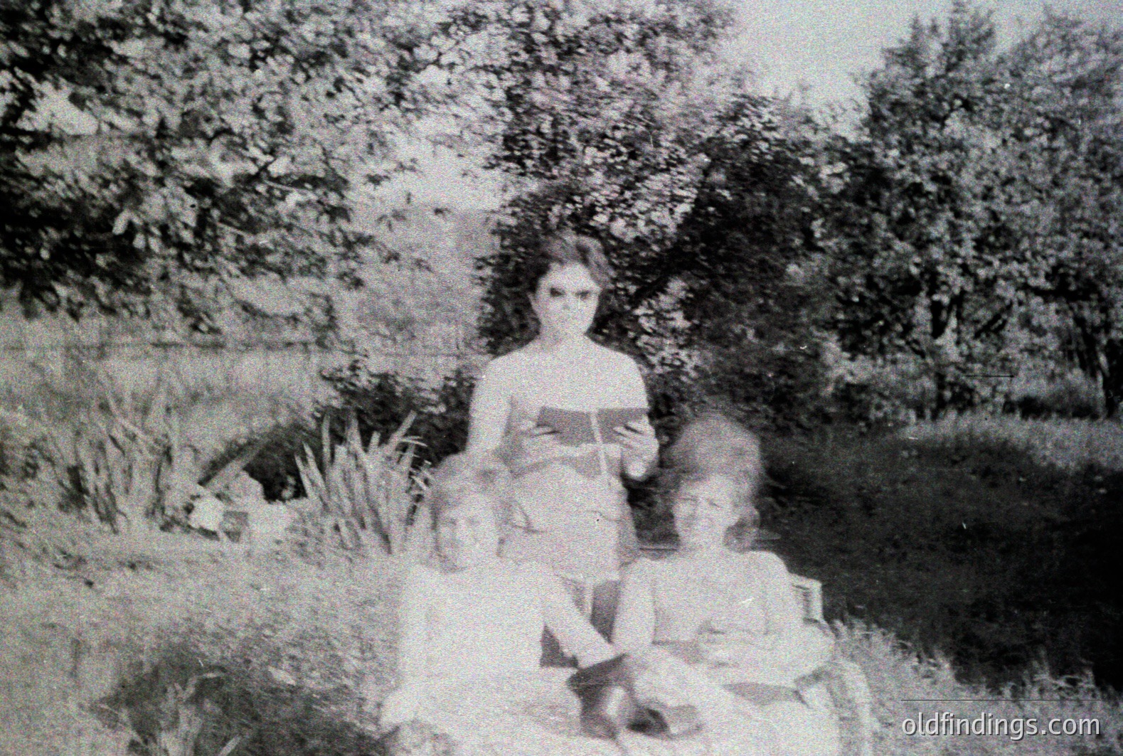 Black and white photograph showing a woman seated with two young children. The woman holds a book, positioned outdoors in a grassy area with dense foliage backdrop. Likely a family portrait, suggesting a candid moment. Appears to be from the early to mid-20th century, possibly 1920s-1940s. Valuable for family history research.
