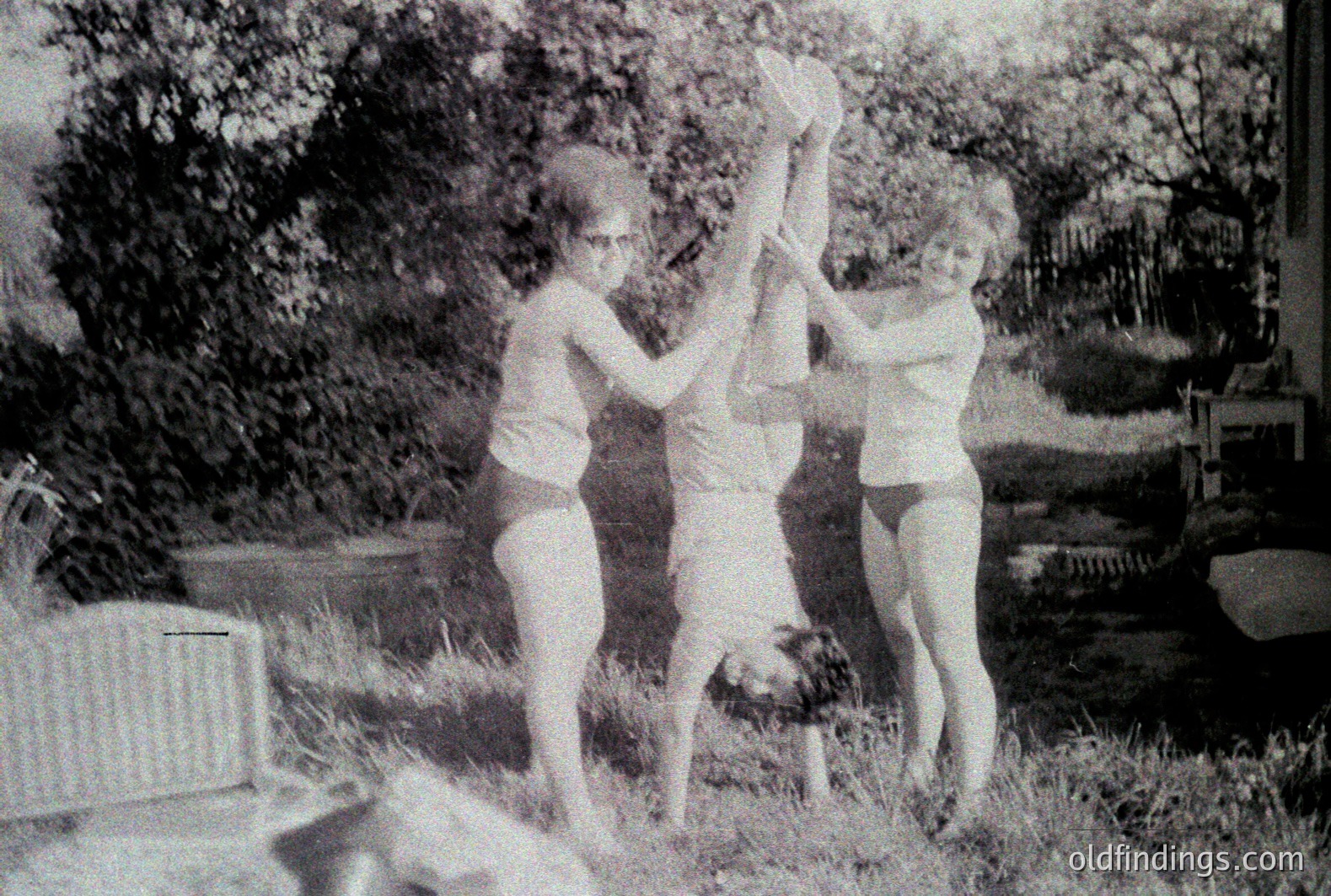 Three young women playfully balance two others upside down, suspended by their ankles. The outdoor scene features a grassy lawn, shrubbery, and a glimpse of a building in the background. Likely a casual snapshot from the 1960s or 1970s, showcasing mid-century leisure. Photographic quality suggests a personal or family album origin.
